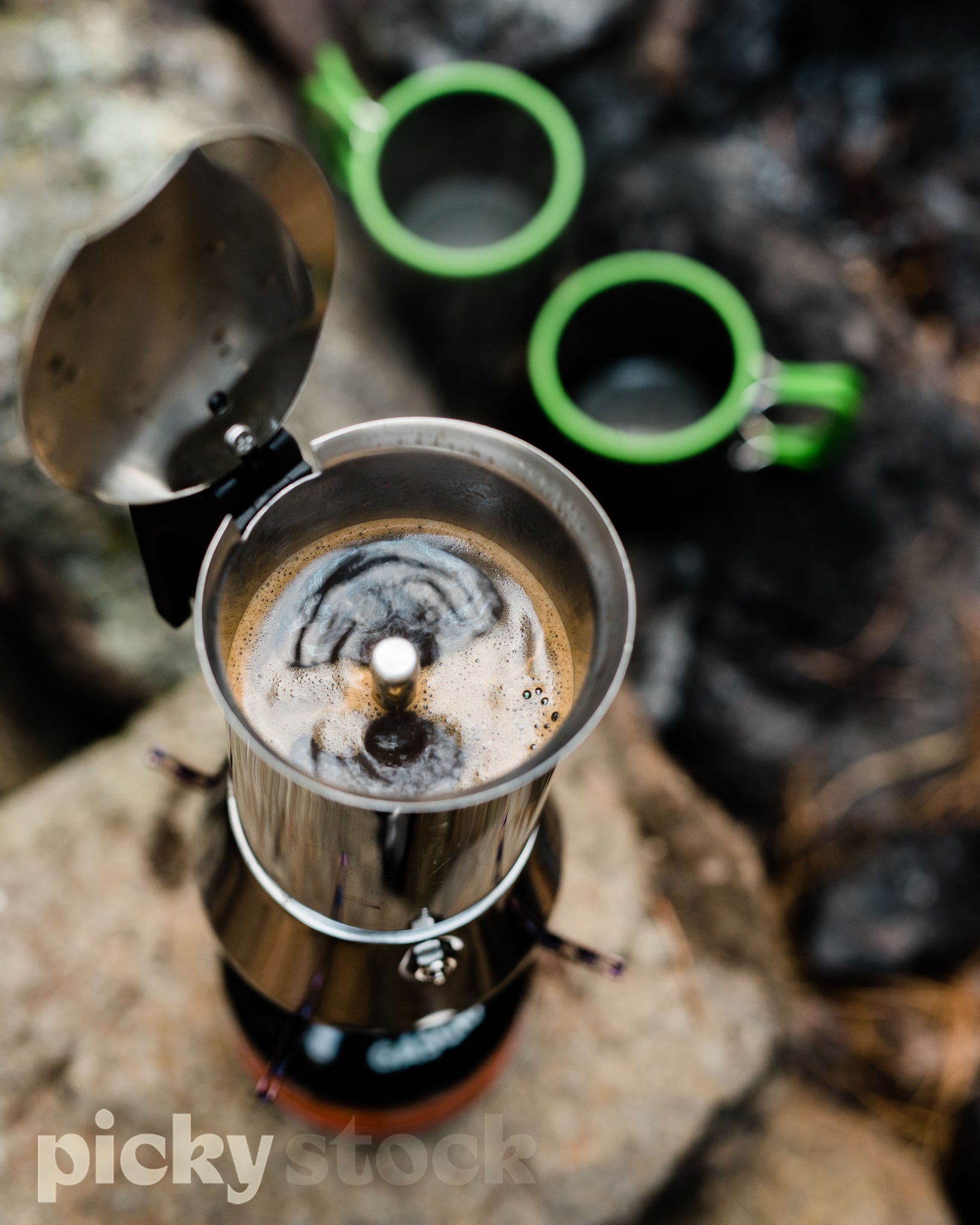 Close up image of coffee maker sitting on a rock. Two empty green and grey mugs in the background ready to be filled. Coffee maker is stainless steel, the burner is black with an orange base
