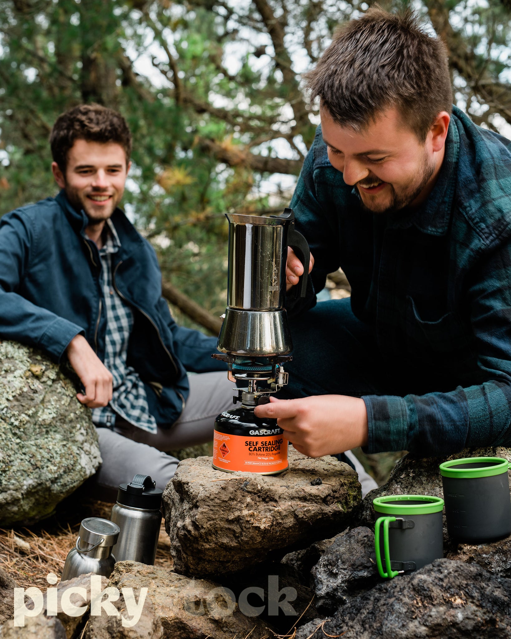 Two friends making a coffee in the outdoors together. One man with brown hair smiling looking down at coffee stainless steel maker. Lighting the gas. Other man also in a blue jacket watching in the background. Two mugs in the foreground right of frame