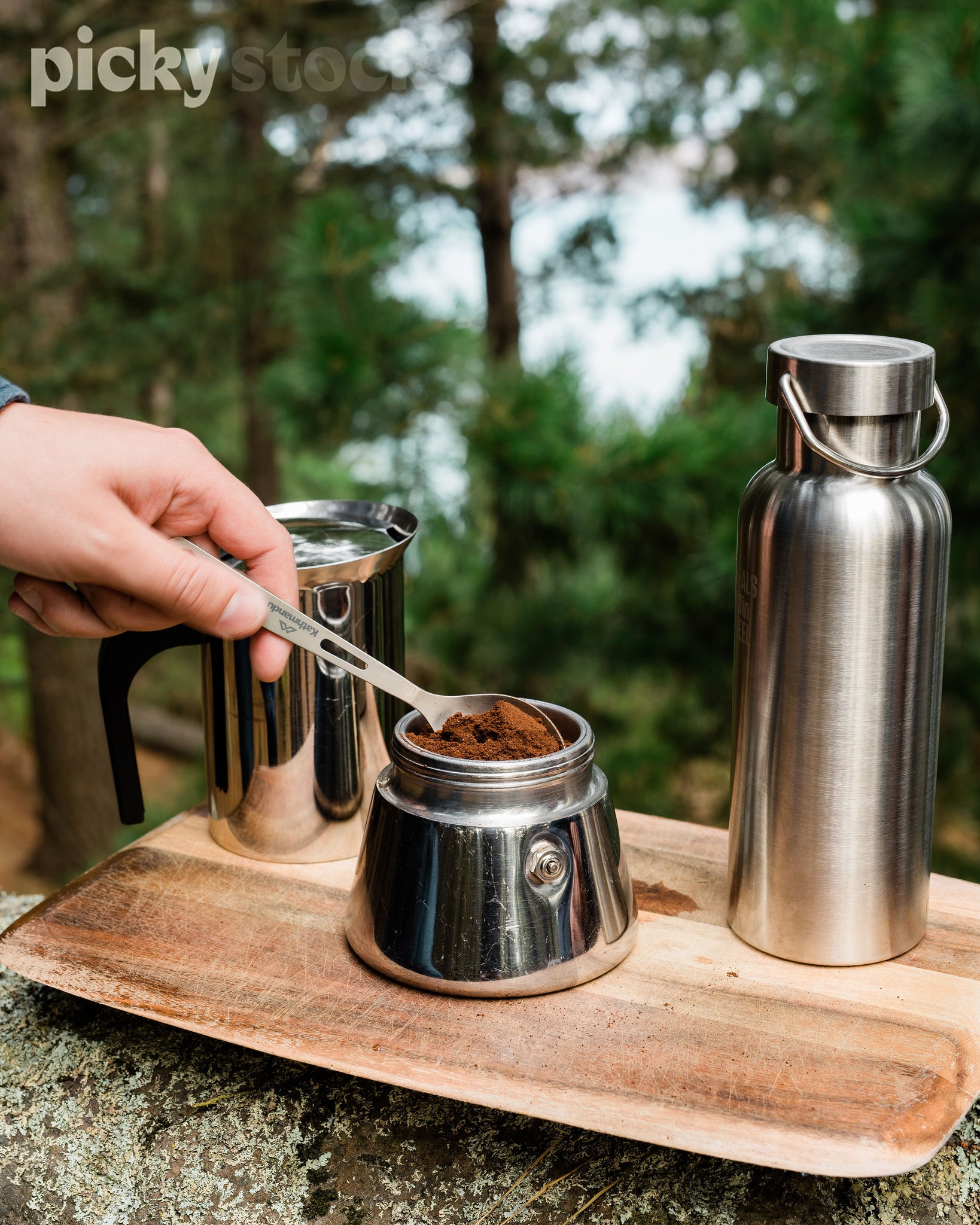 Man scooping coffee portion from jar onto spoon. Coffee container, stainless steel water bottle and jug sitting on top of wooden chopping board, on top of a log. Background is trees and greenery. 