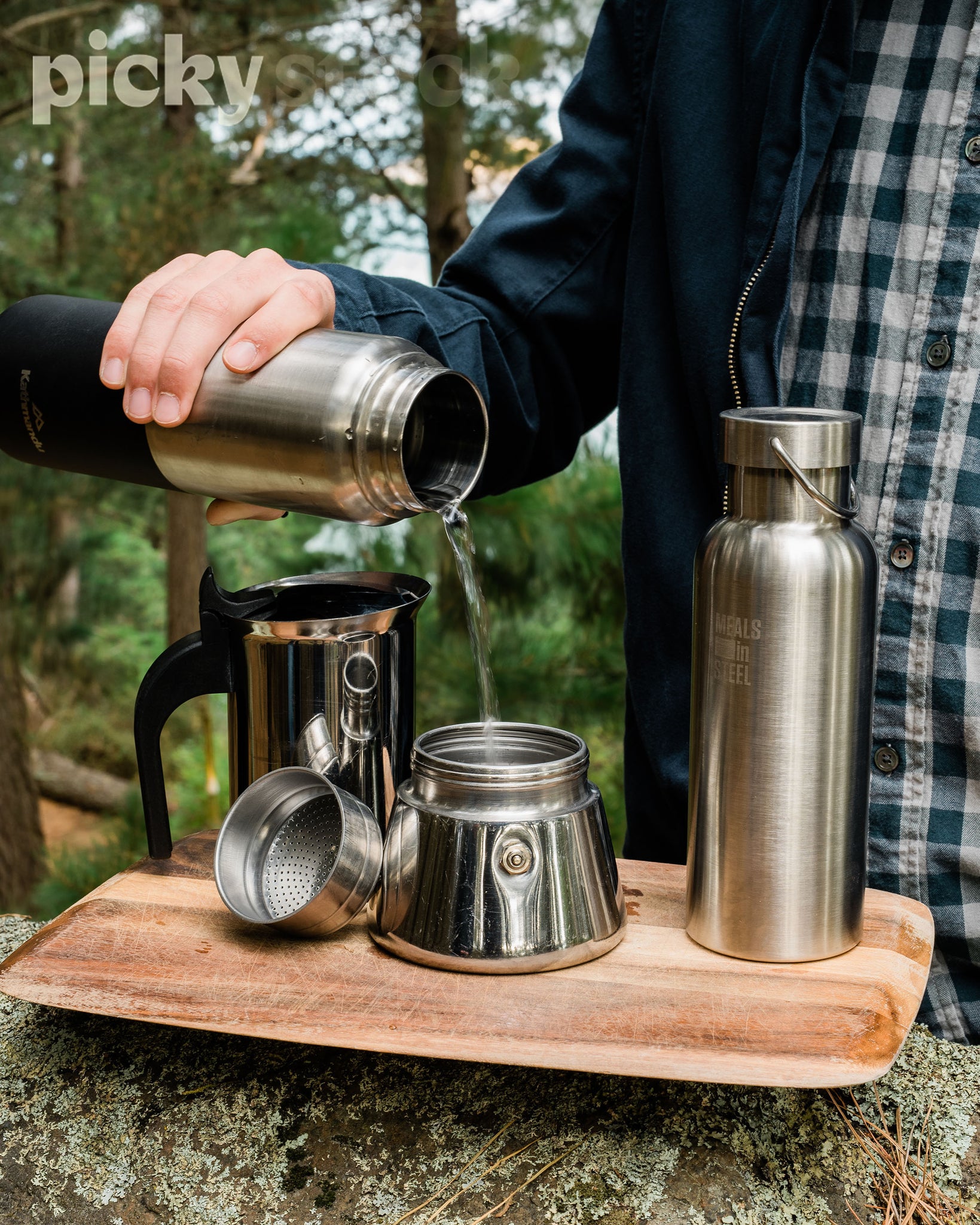 Man pouring water from stainless steel thermos into cooker to make coffee. Two grey mugs with green handles in the foreground. Man is wearing a blue jacket and light grey pants. Coffee maker, stainless steel water bottle is sitting on top of wooden chopping board, balancing on a log. Green trees and other greenery in background. 