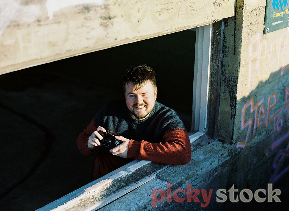 Gay Man wearing red and navy jumper looks out window of run-down building holding 35mm Film Camera