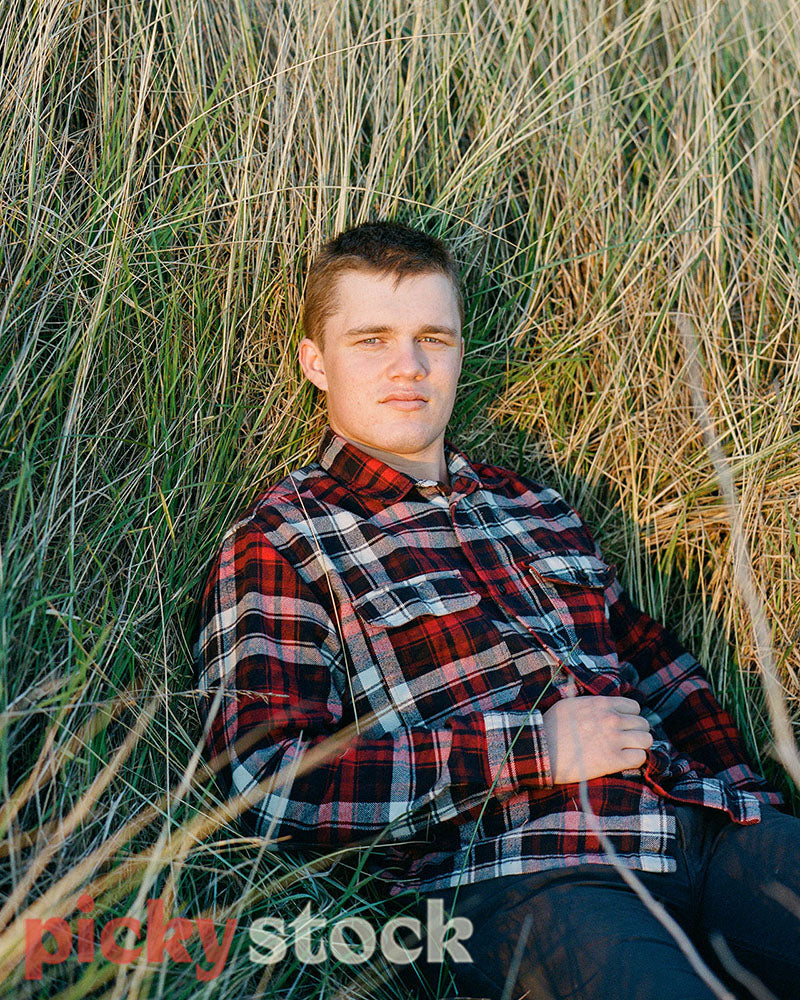 Gay Man laying on tussock grass, while wearing a check shirt., gazing to camera. 