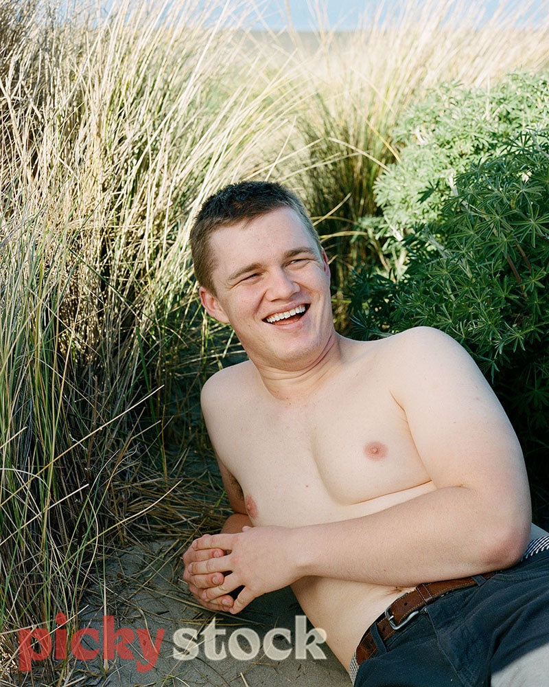 Gay Man laying on a sandy beach, near the grass, while shirtless. He gazes just off camera smiling.