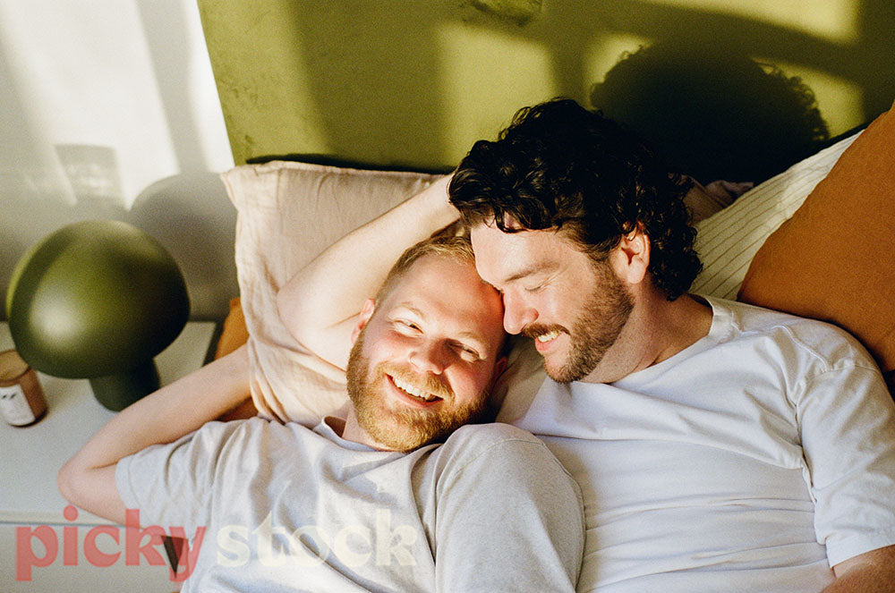 Gay couple cuddling on a bed, wearing white tee shirts. Green headboard