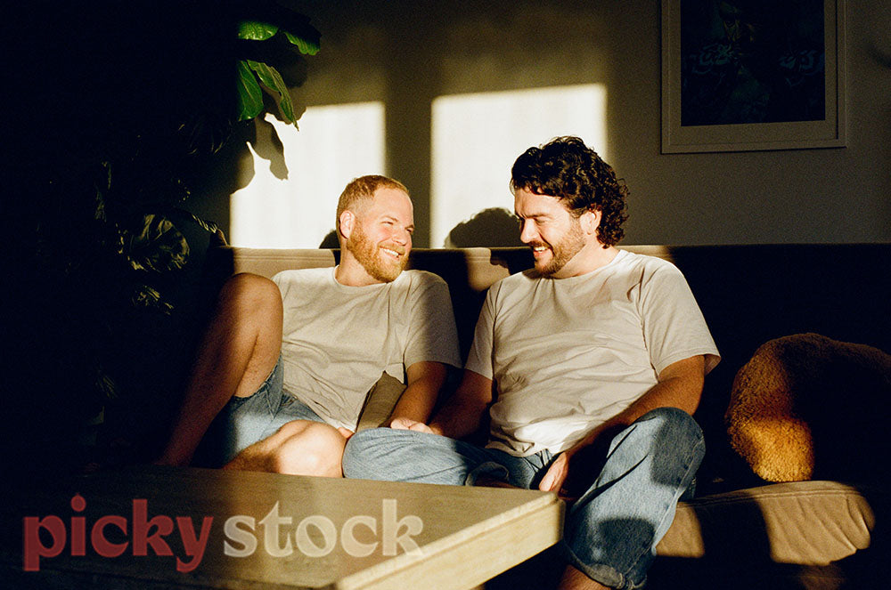 Gay couple sitting on couch looking at each other smiling. 