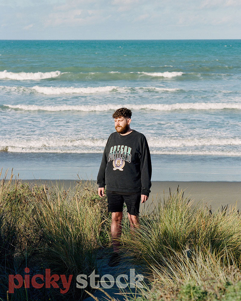 Gay man standing on a sand dune, at beach. The rolling waves  behind him as he looks back to shore.