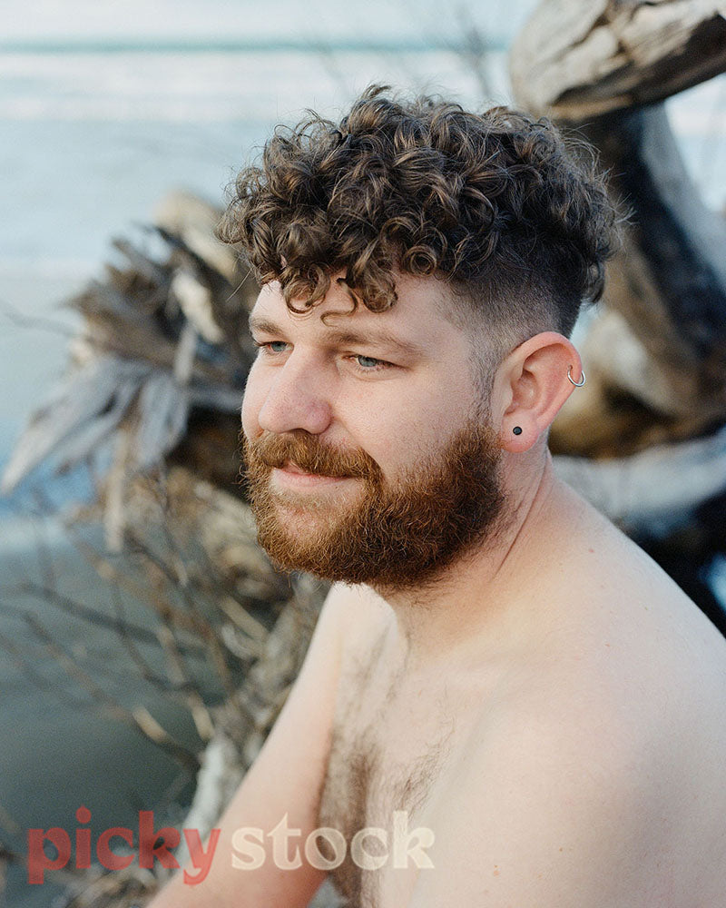 Close up of shirtless gay man sitting on a washed up tree at the beach