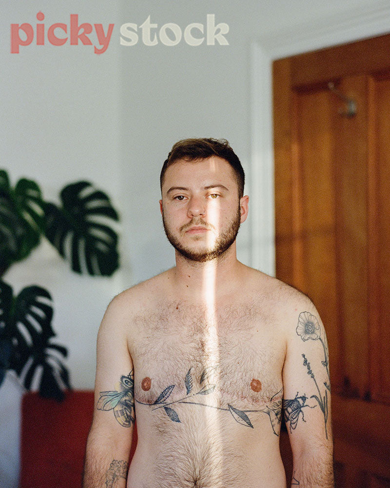 Trangender man portrait, taken in a bedroom. Not wearing a top, tattoo's on body.