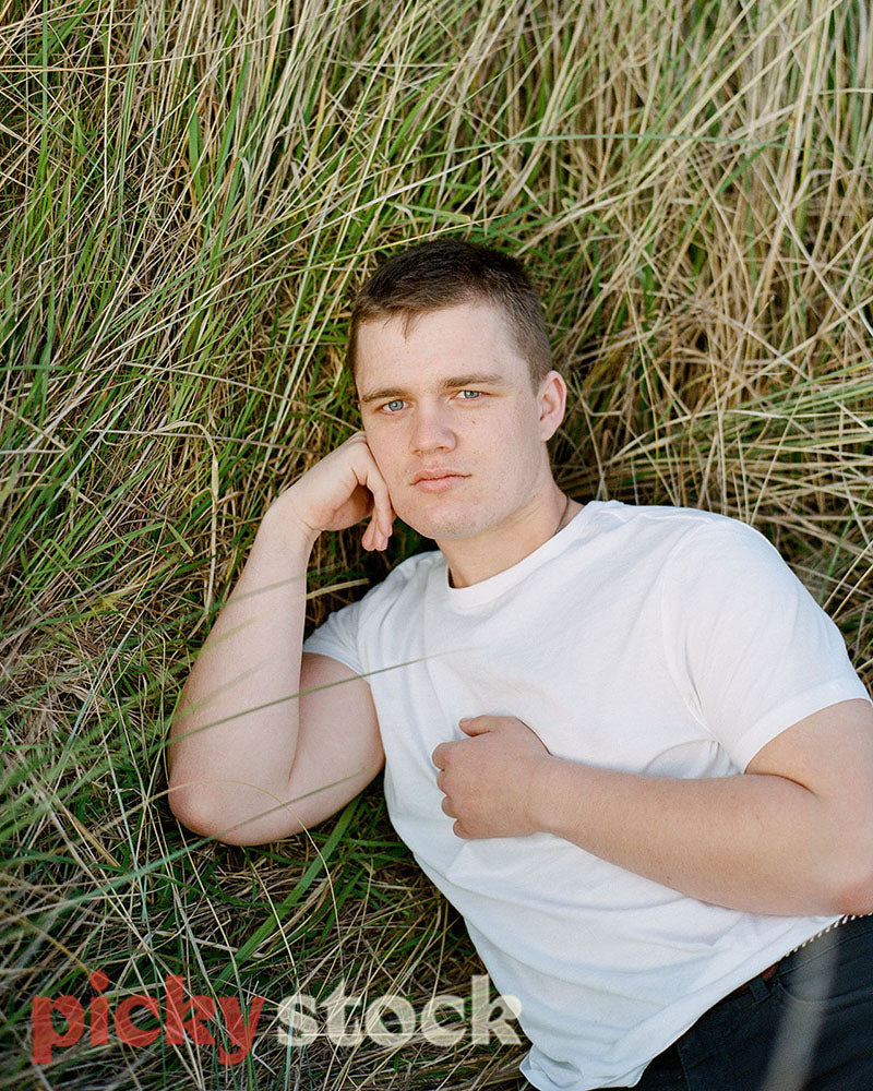 Gay Man wearing simple white tshirt laying on a tussock beach, looking to camera.