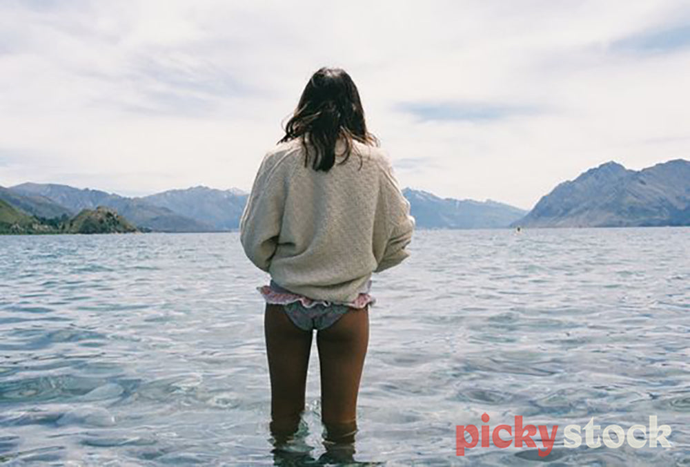 Lady stands in waters of Lake Hāwea, tog bottoms and a jumper on.