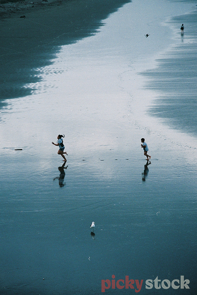 Two kids playing tag on the beach, amongst the glistening sand, seen from afar. 