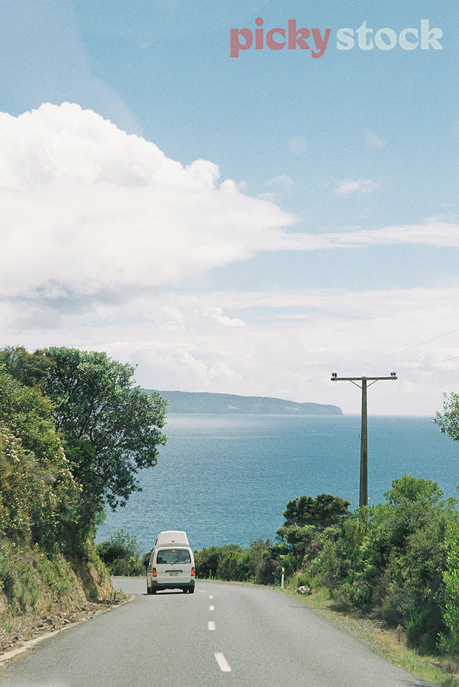 Van on the open road, up hill looking out to blue ocean on a blue day.