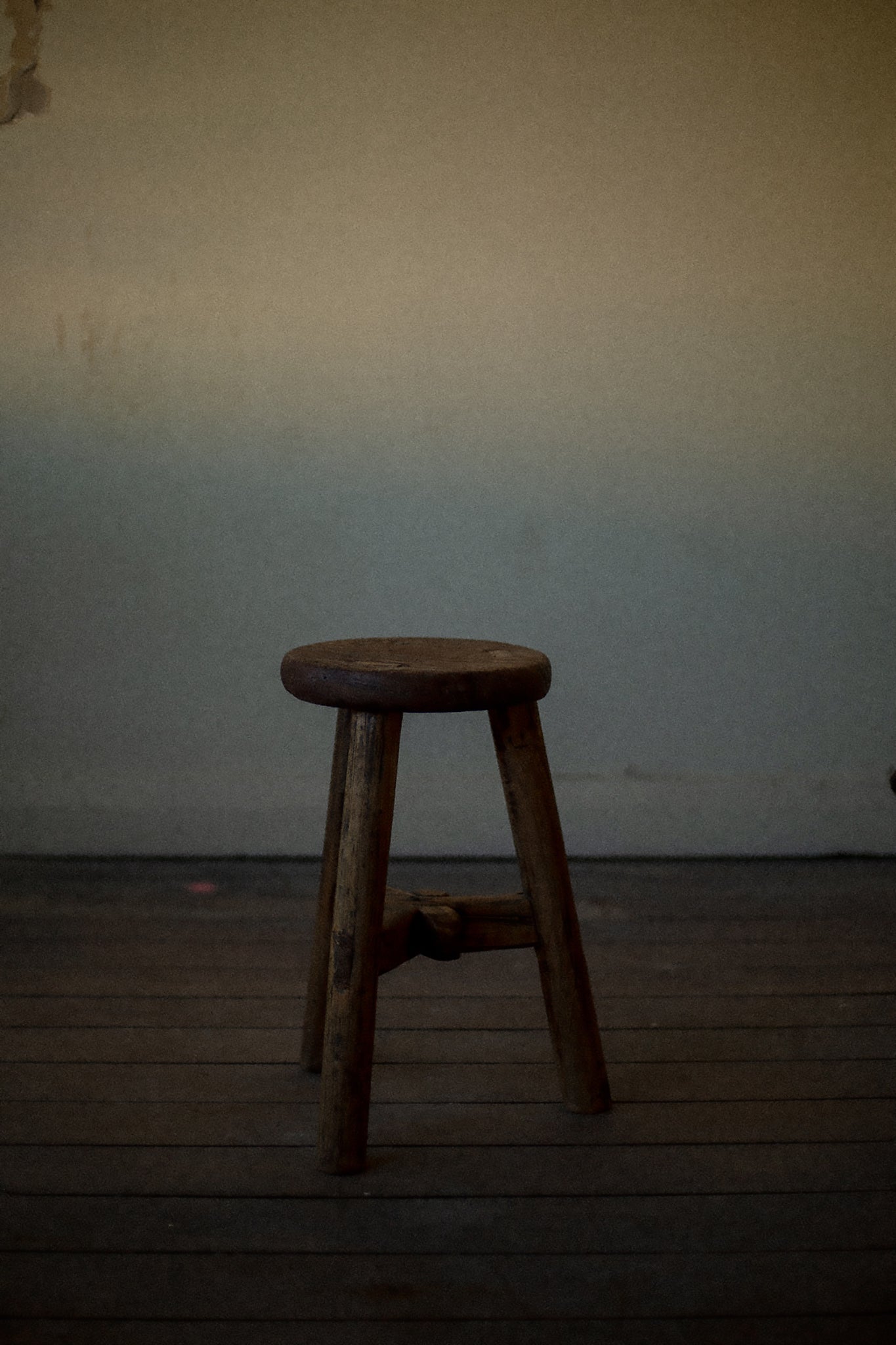 A little wooden circular stool on hard wood floor, the light is gloomy as day comes to a close.