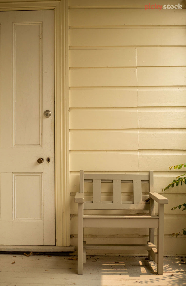 Charming front porch where a one-seater wooden chair sits against cream weatherboards beside a cream door. 