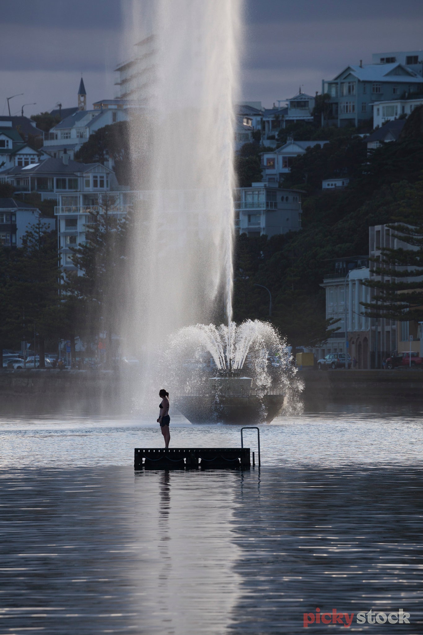 Single girl in swimwear standing on the pontoon at Oriental Bay. Large water feature behind her with houses on the cliffs along the waters edge. 