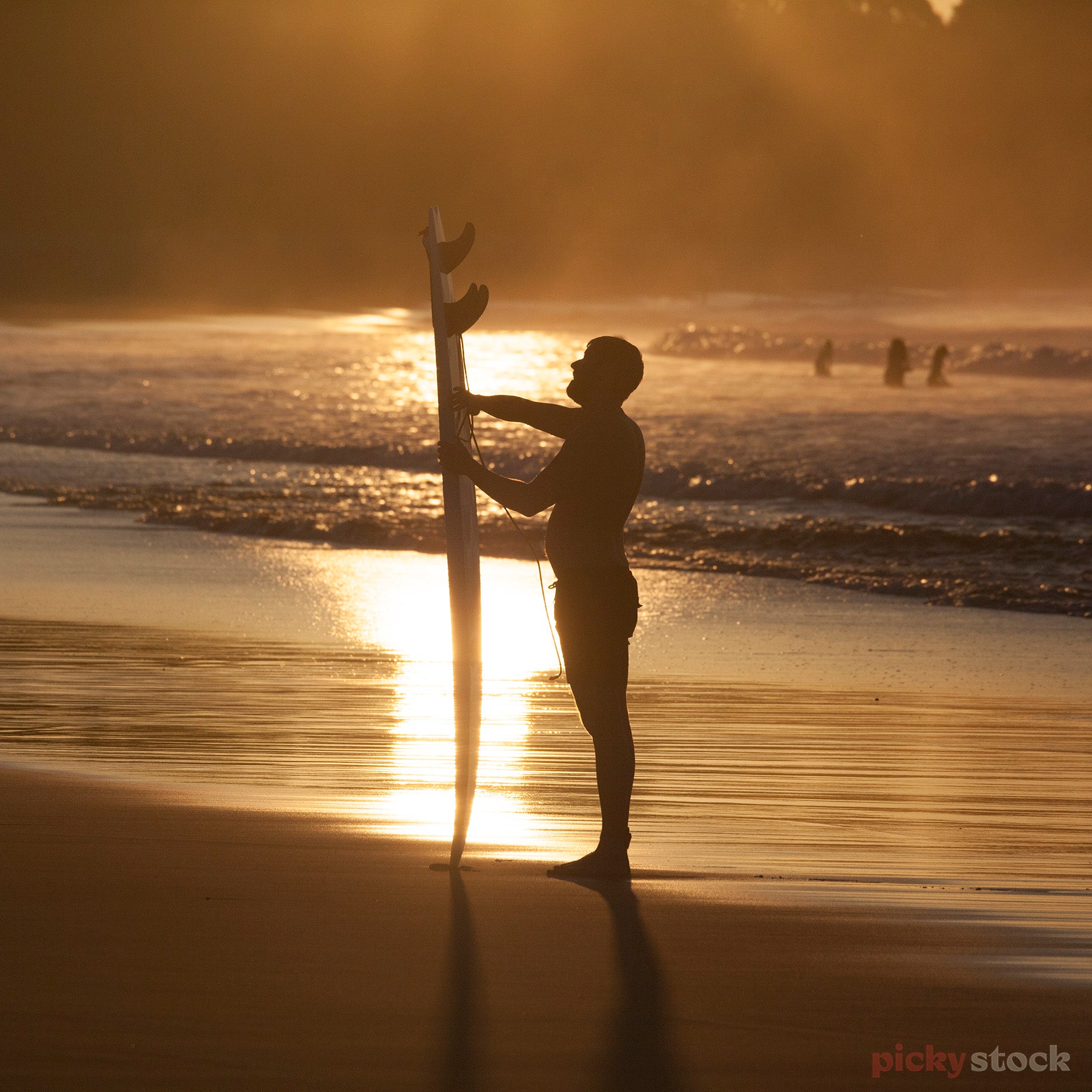 Surfers at Oakura Beach at sunset / sunrise. One man standing close to camera looking up to vertical surfboard. Surfers out in the ocean n the distance. Sky is golden,. 