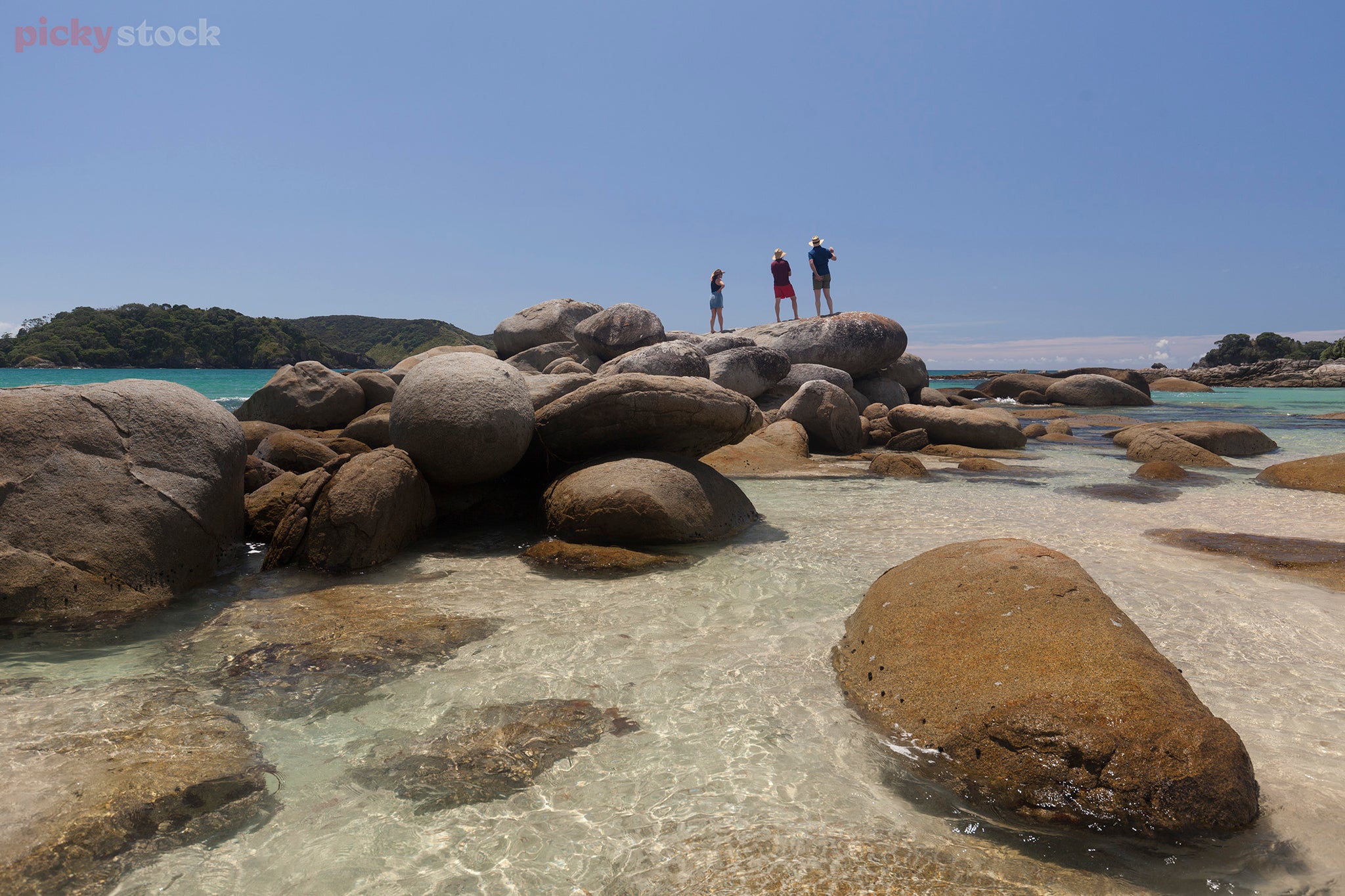 Three people standing on the rocks at Matai Bay.  Bright blue sky with large rocks and clear water. Golden sand visible. 