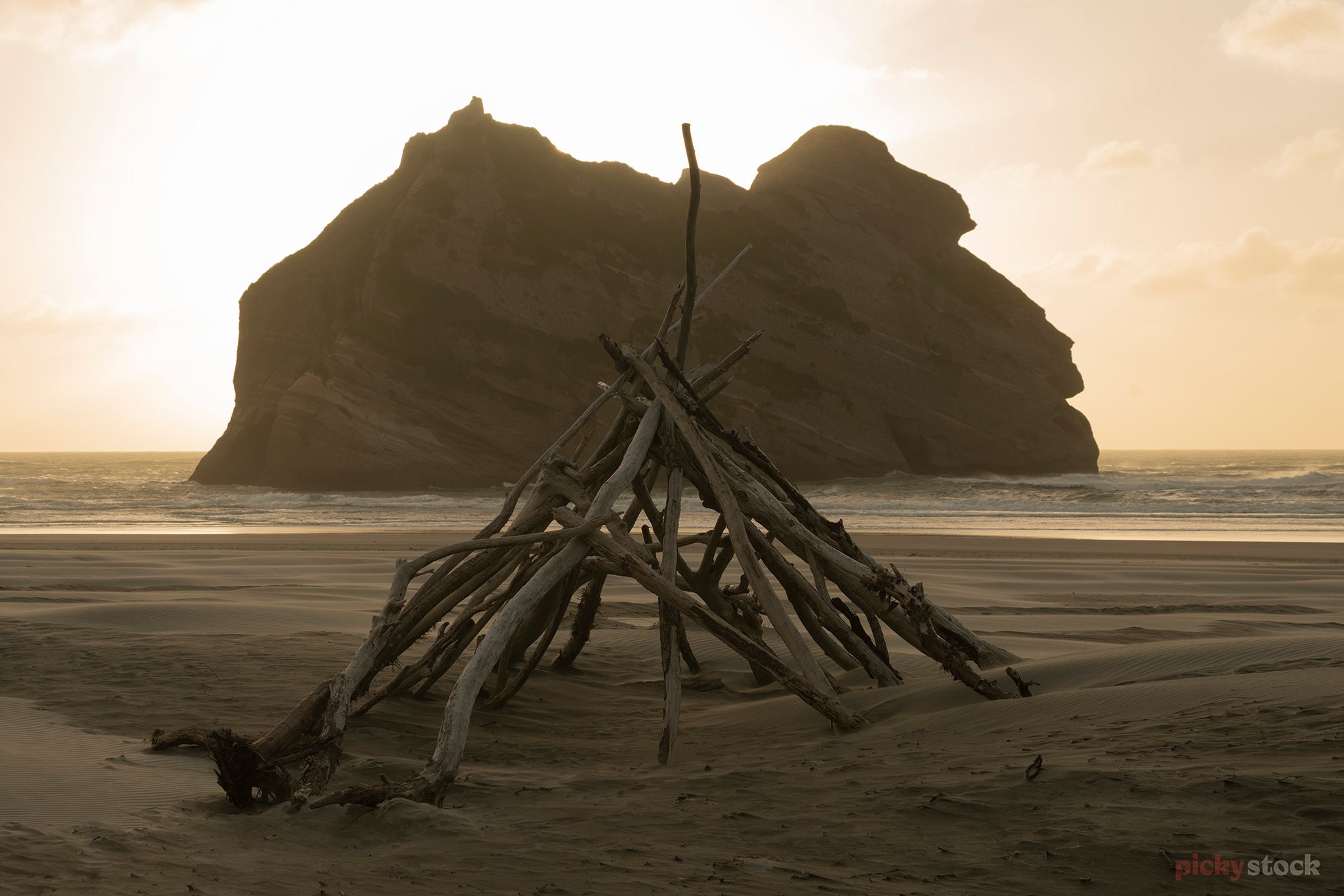 Driftwood structure tipi at Wharariki beach. Golden light on sand beach. Large rock formation in background. 
