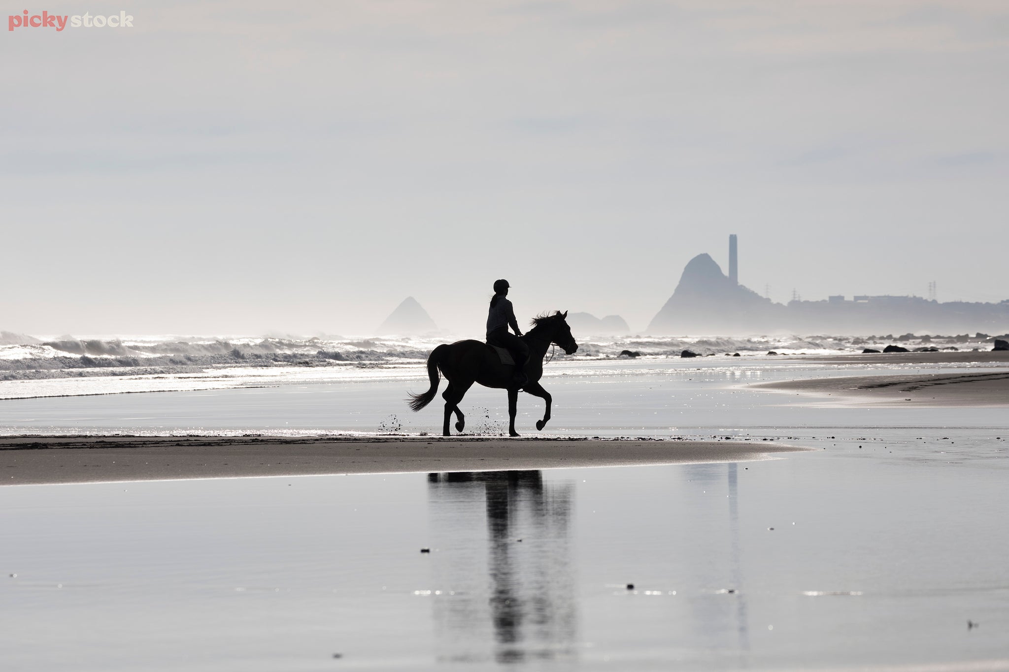 Lady riding a horse along a beach in Taranaki. Soft grey and blue sky. Lady is in shadow - dark outline. 