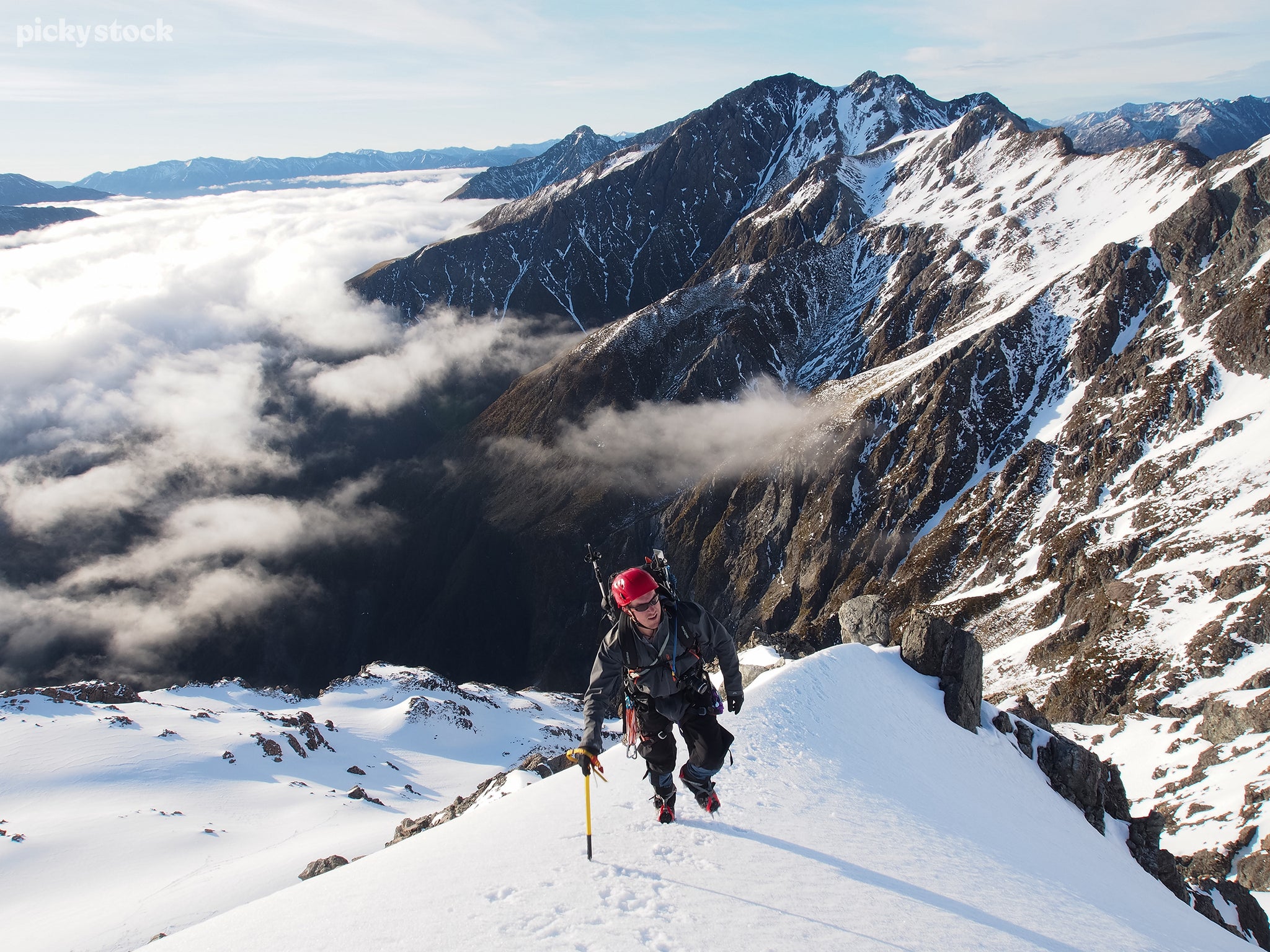 Landscape of man climbing a snow-capped mountain higher than the clouds. the climber advances the trail with a yellow walking stick. The background reveals an endless valley of peaks.