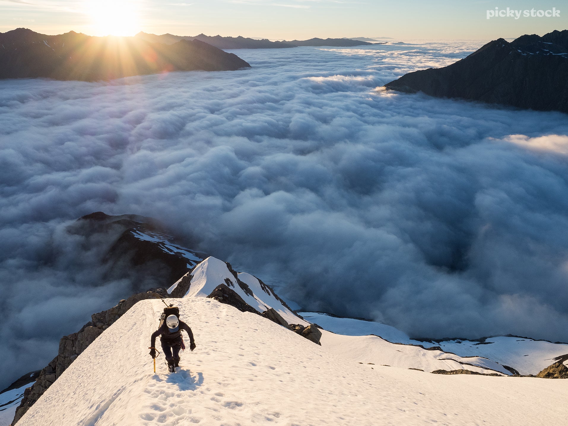 Landscape of a woman climbing a snowy mountainside, the climber advances steeply up a sharp incline well above the clouds. The sun climbs over the jagged peaks in the background, casting a warm field ahead of the climber.