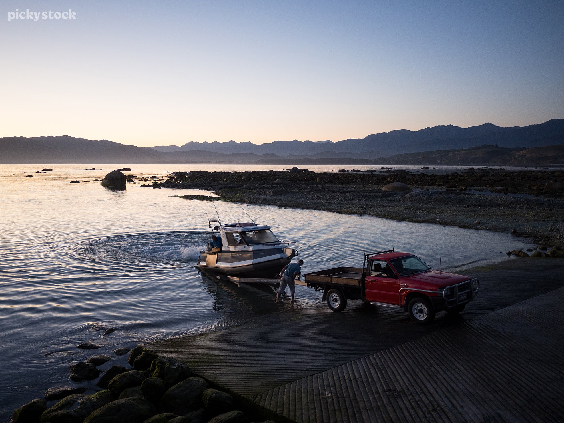 The landscape of a group of men loads a boat onto a trailer from the water's edge while one ma operates the wench. The boat trailer is attached to a red ute parked on a concrete jetty.