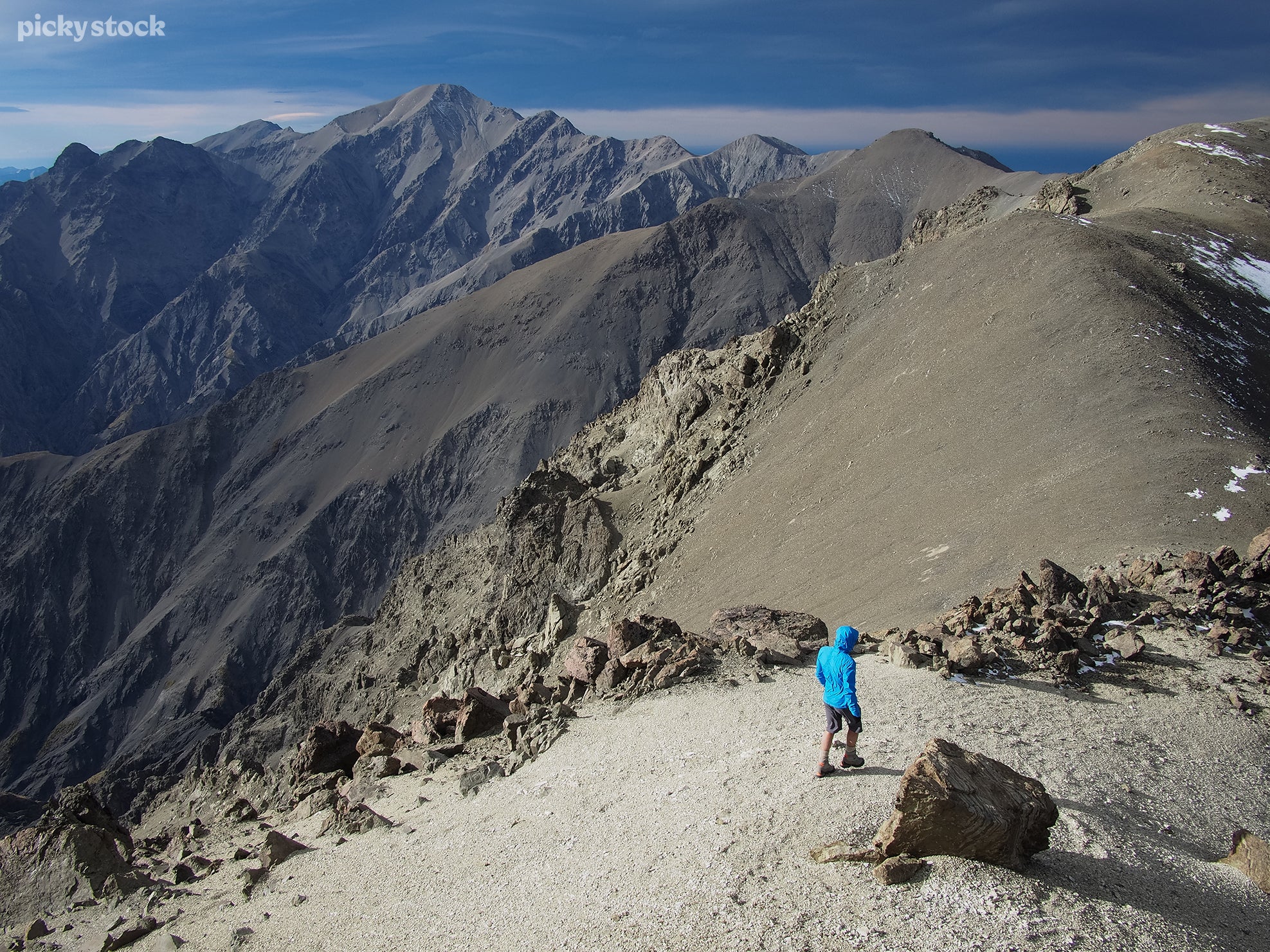 The landscape of a man in a bright blue jacket walks down a path descending from a rocky mountain side. A dark blue sky looms high above the trail.