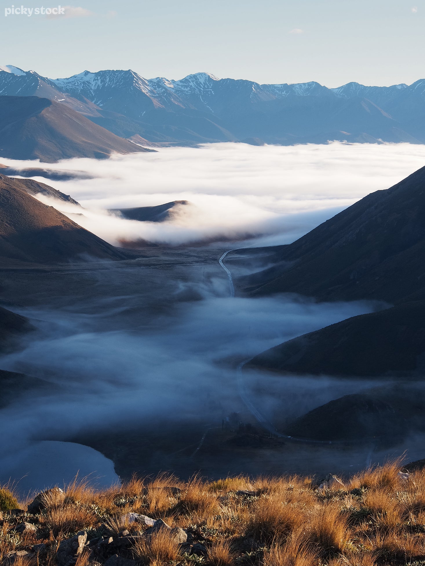 Portrait of a mountainous valley from atop a hill of rough brown grass. A single road winds the shady contours of the valley and disappears into a lake of dense white fog.
