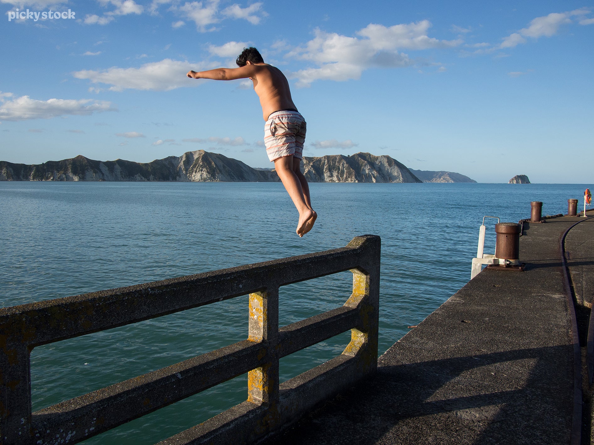A landscape of a man in swimming trunks leaps into the water from a concrete dock. The sunny sky casts light across the water reflecting off brass cleats and white cliffs.
