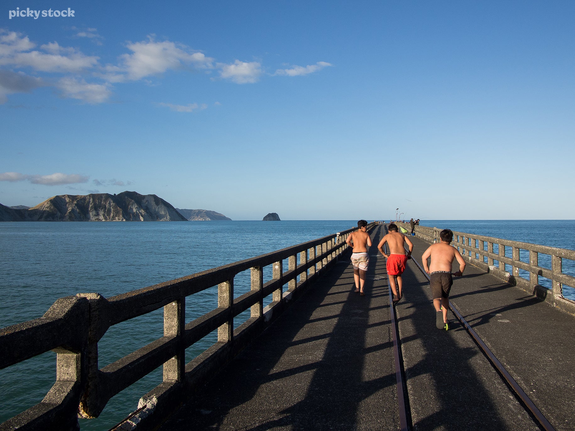 A landscape of three young people running down a concrete jetty towards the water. The sky is bright and shines dull against the concrete handrails and white cliffs.