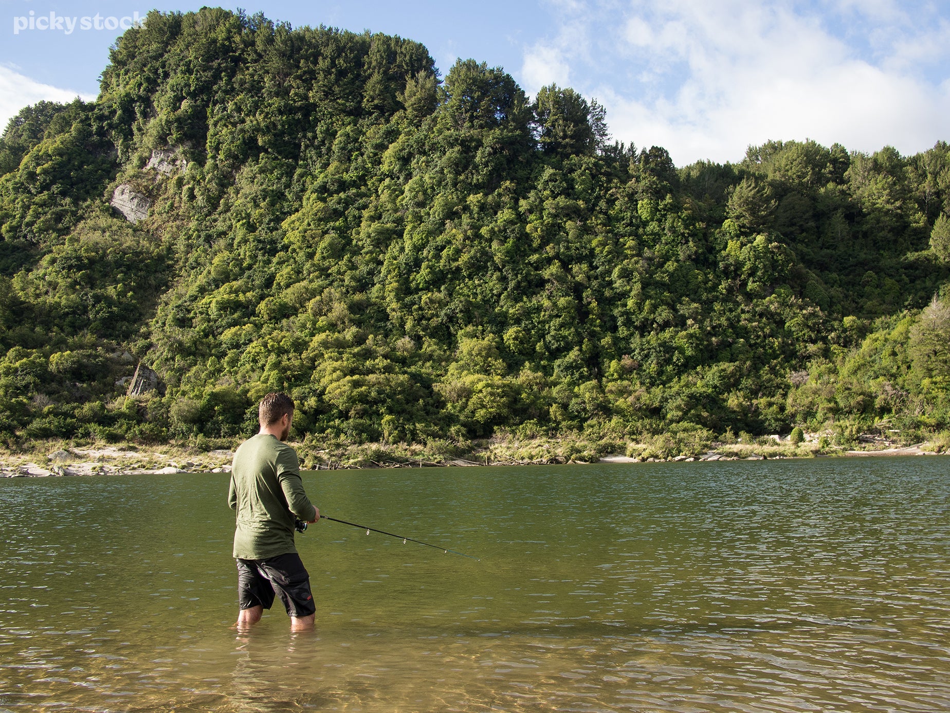 A landscape of a man fishing in a wild stream. His dark shorts are drenched with water and his fishing pole is angled down in anticipation. The water is murky green and reflex the bevy of trees that coat the hillside.