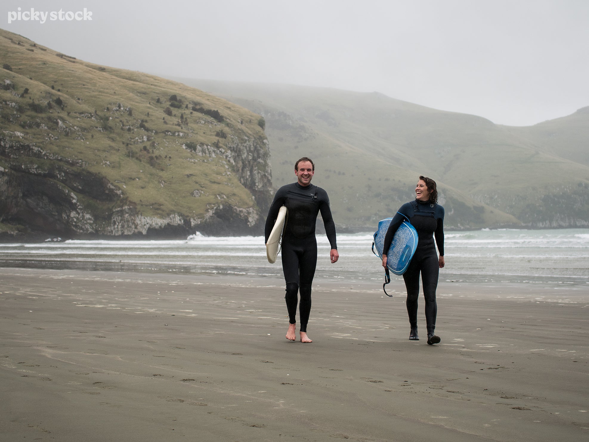 Man and woman walking holding surfboards along high tide line. Foggy low cloud day. Early morning or early evening. Black sand beach. 