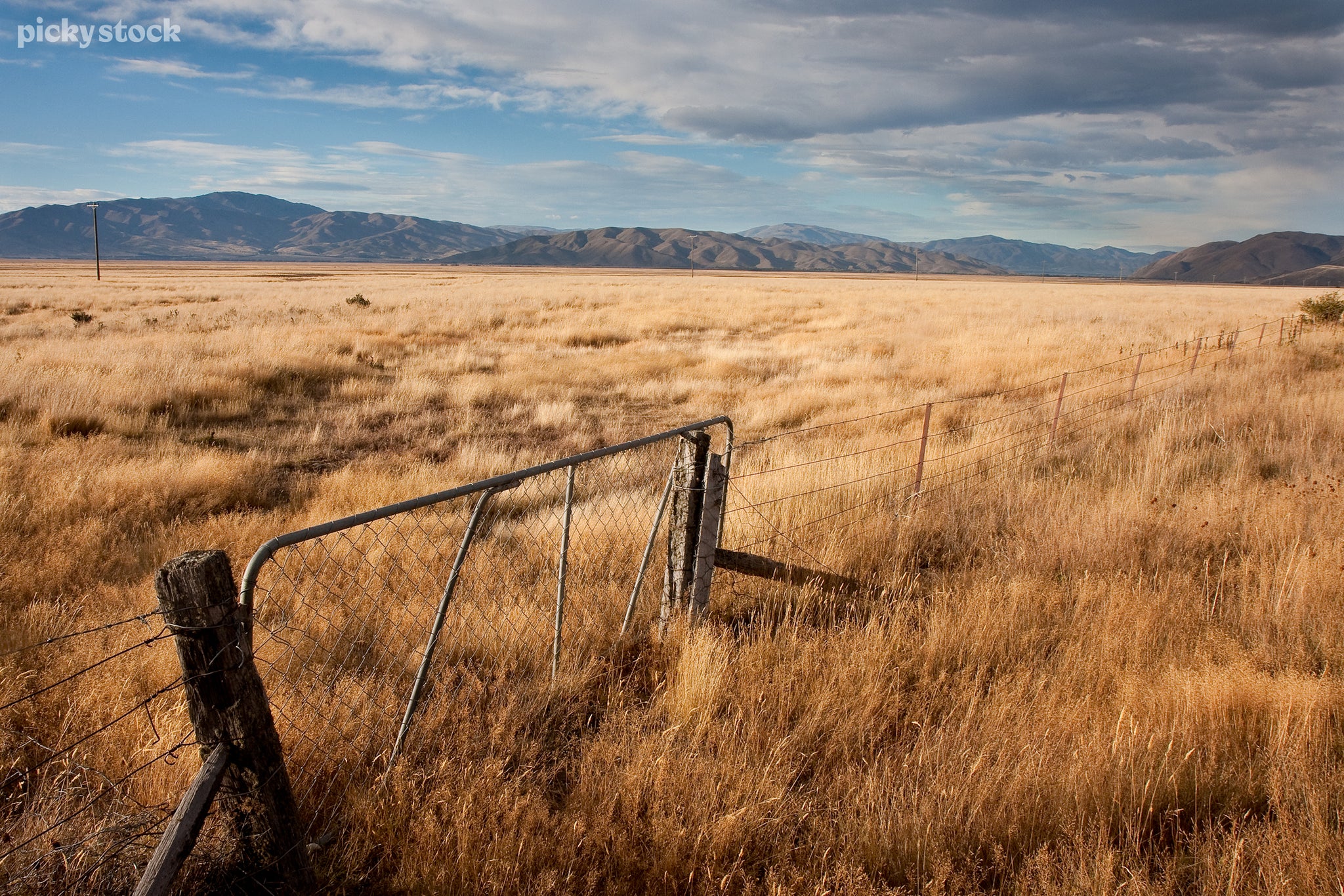 A landscape of an old wire fence and iron gate blocked access to a lush plain of golden grass. The flatlands stretch far towards a mountainous range while clouds list atop the fields.