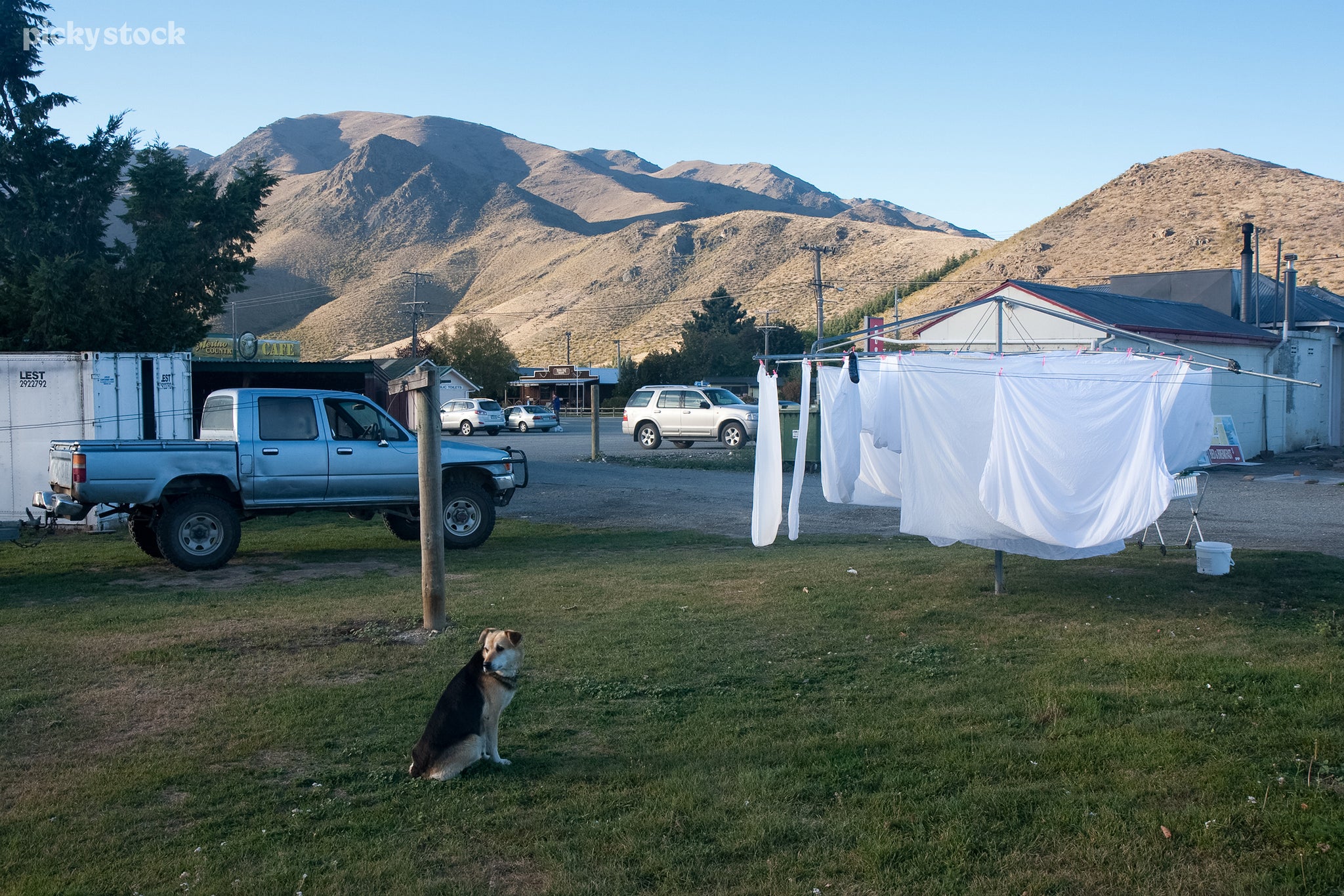 A landscape of a small cluster of buildings in between green hills. A medium-sized dog sits patiently next to a blue four-wheel ute while white linens dry outside a travelers motel.