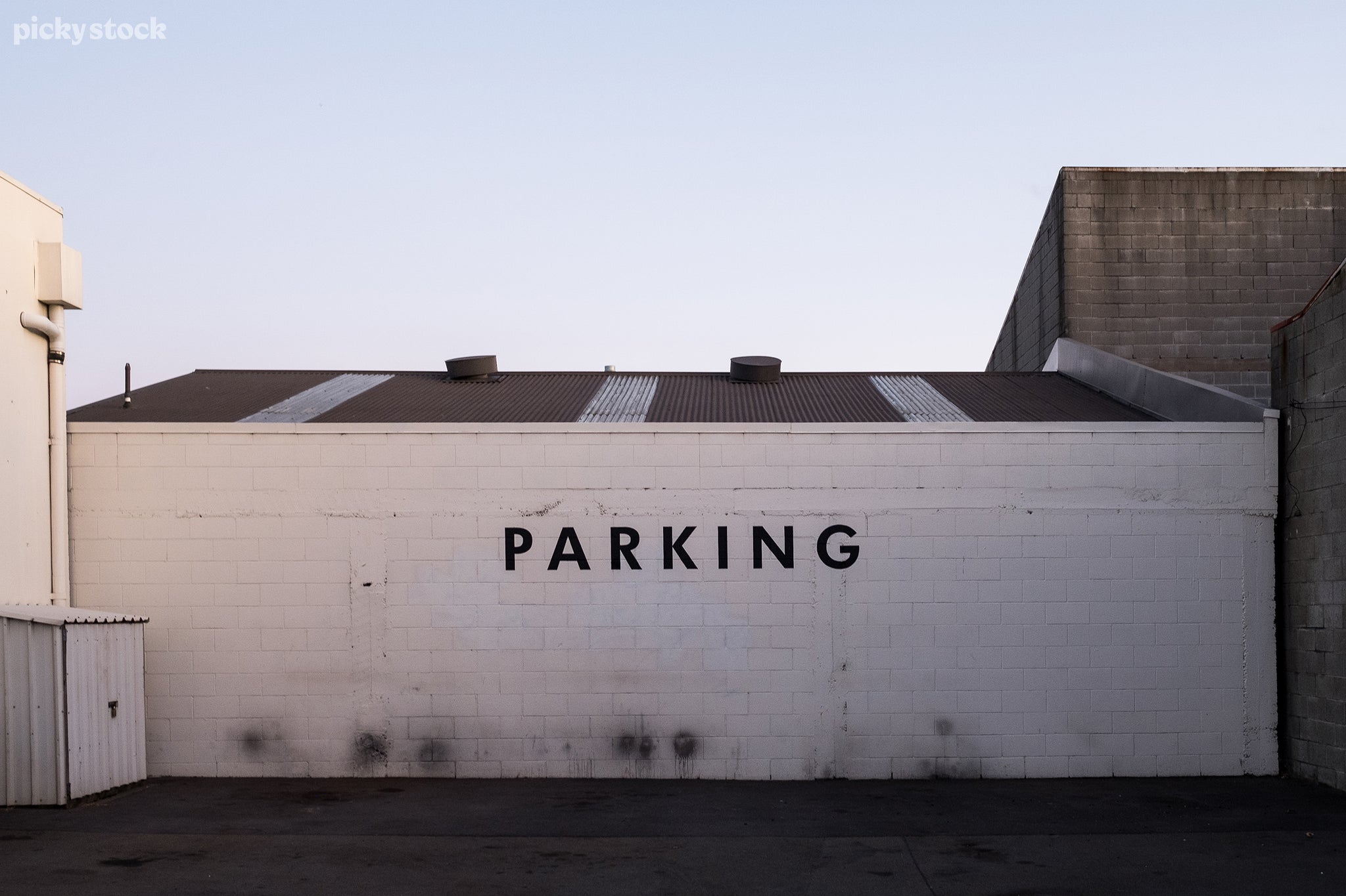 Landscape of a white brick wall with large block letters reading ‘parking’ in spaced lettering, the walls if caked with dirt and exhaust from backfire. The surrounding buildings of grey brick block the light and cast a cool light over the dark asphalt.