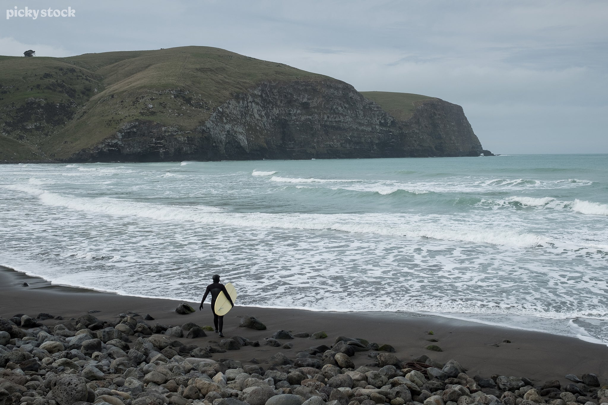 Landscape of a rocky beach and green hillside, a surfer in a black wetsuit and white surfboard looks out towards the water as waves push towards the shore, white crests breaking on contact with one another.