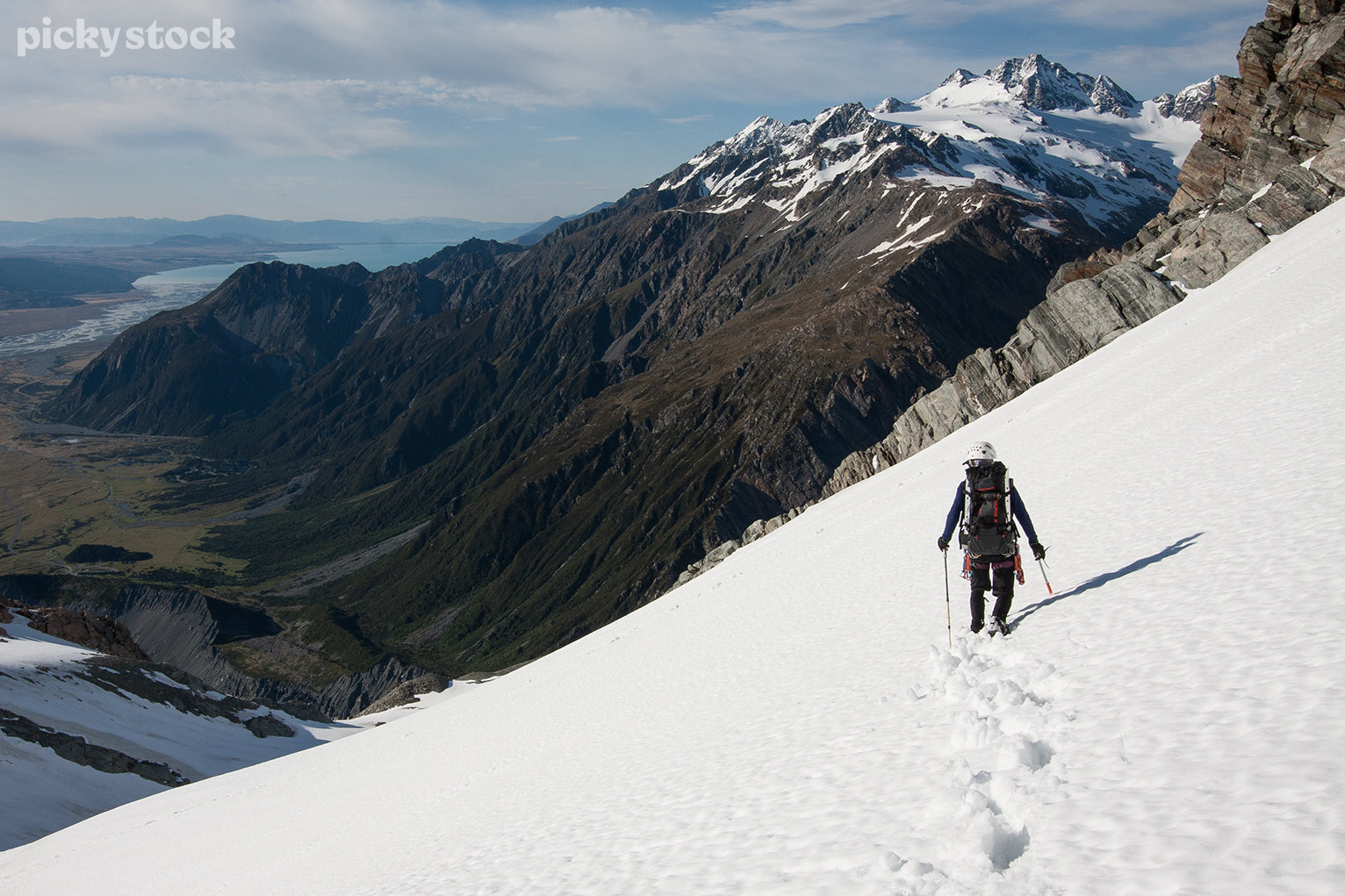 Landscape of a mountain climber with a white climbing helmet and backpack traversing a snowy slope, a deep trail forms behind them. The climber looks outwards to a green valley and river below them.