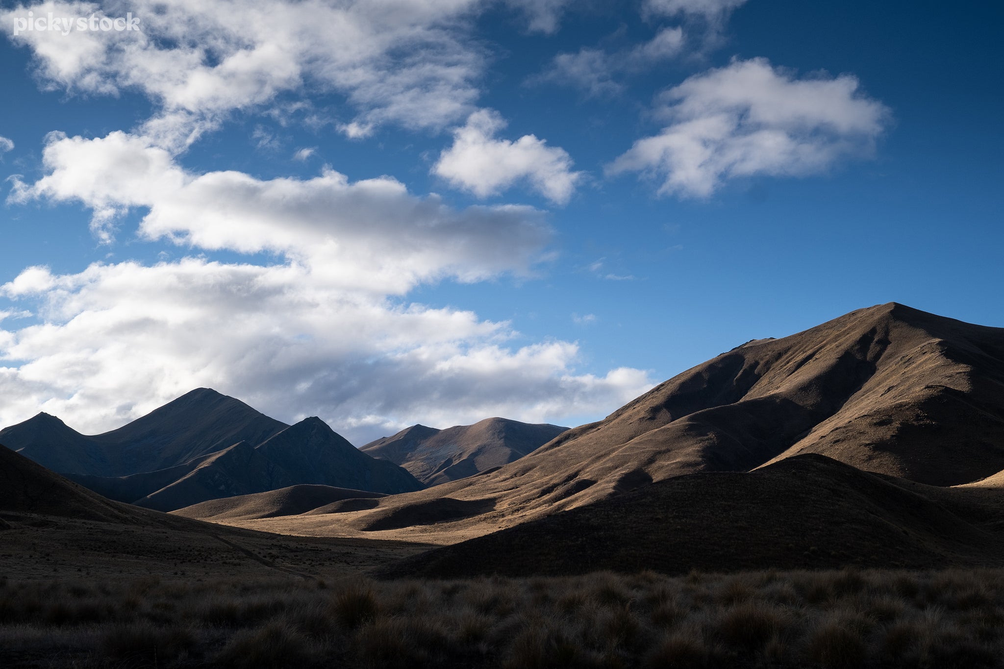 Landscape of brown grass hills expanding across a field. Deep blue sky and clouds float idly past casting long deep shadows across the landscape, their contours creating deep craves in-between the hillsides.