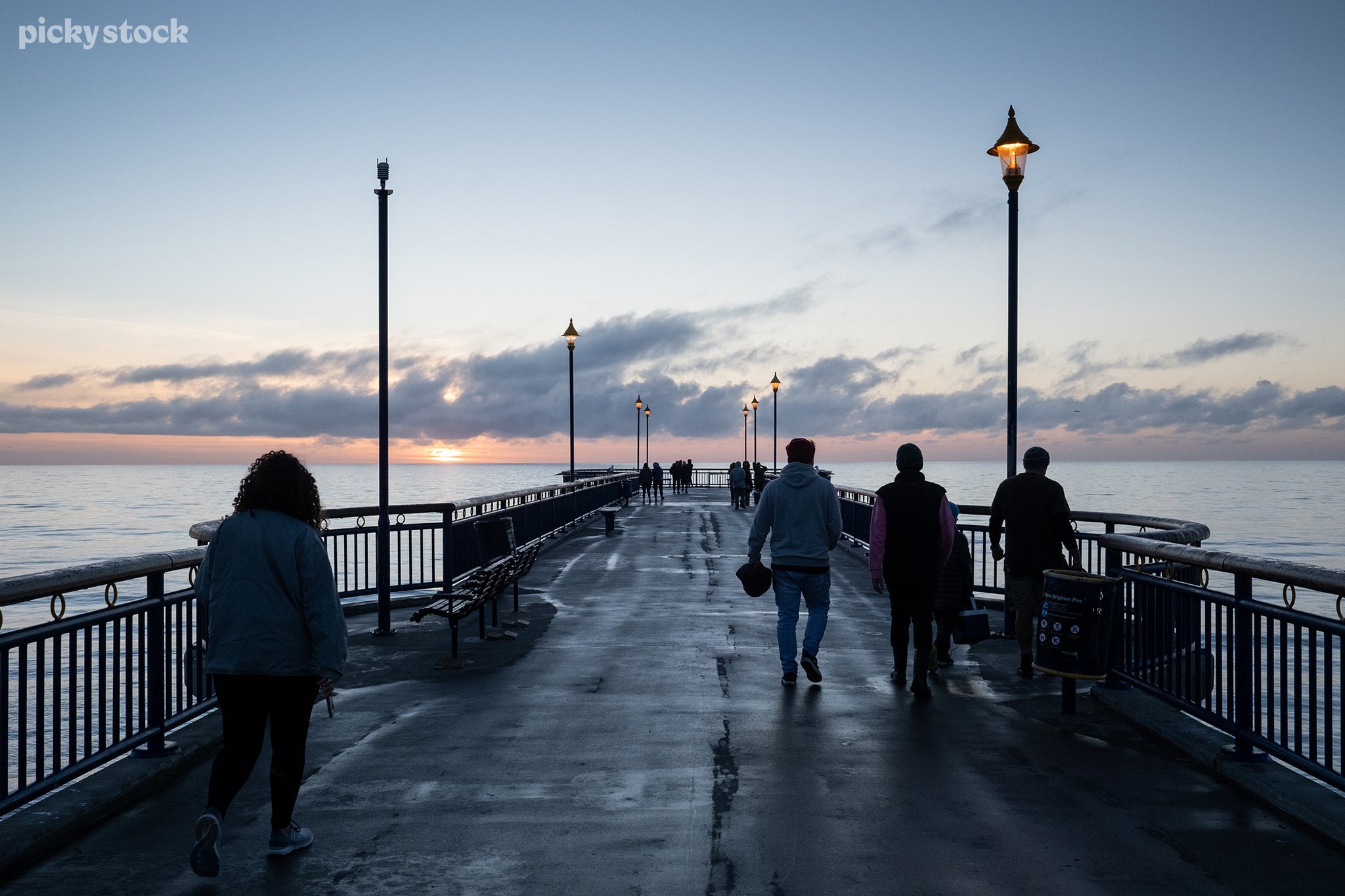 Landscape of a wet concrete pier in the afternoon, tall lanterns cast a soft orange glow on the people below. The sun sets in the distance as many sightseers amble towards the end to watch its final descent.
