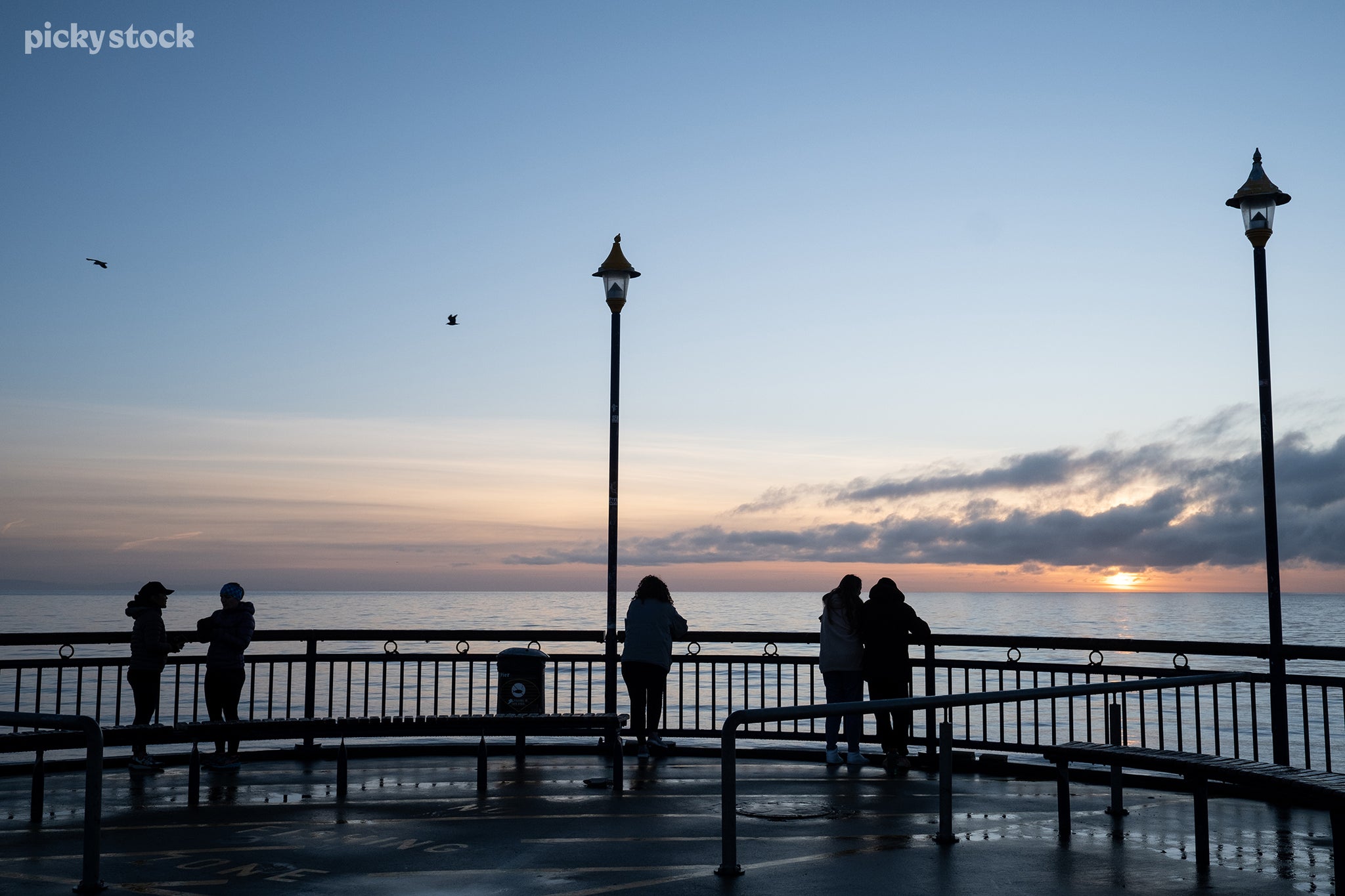 Landscape of people on a wet concrete pier in the afternoon, tall lanterns stand extinguished. Sea birds loft about and clouds crawl across the water as the sun makes its descent.