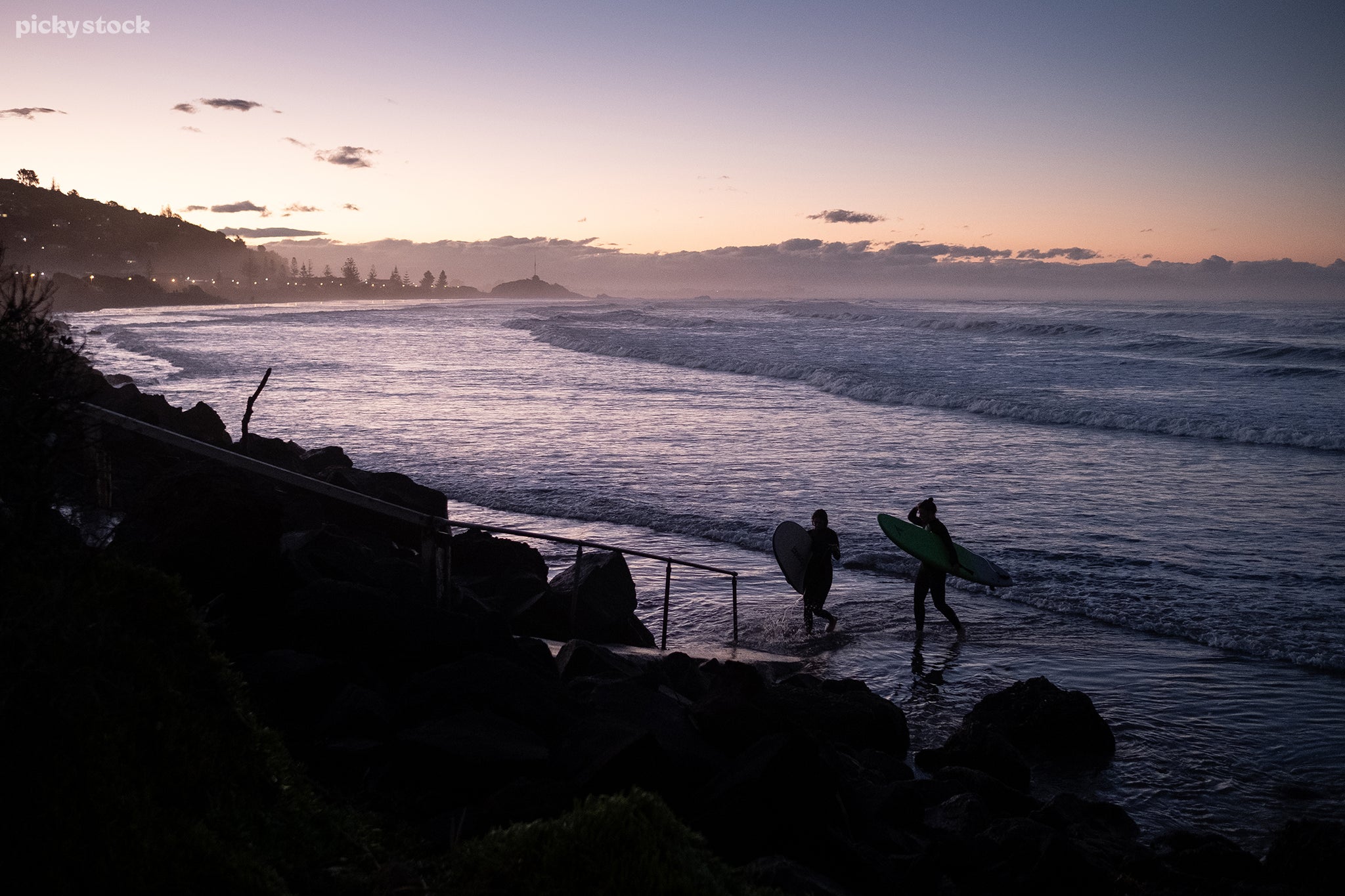 A landscape of two female surfers in black wetsuits leaving the water for a concrete ramp built int a rocky flood wall. The sun sets behind dense clouds while street light glow in the distance.