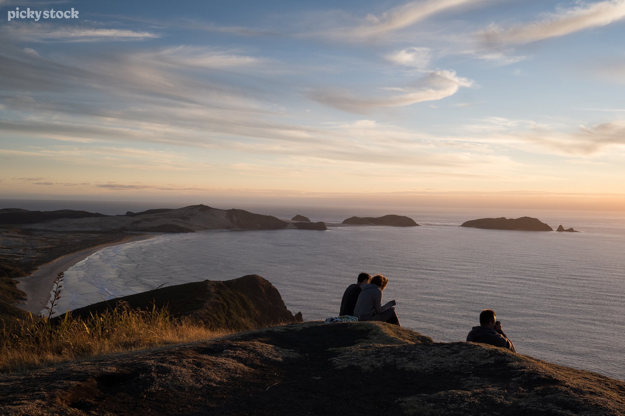 A landscape of three people sitting atop a tall grassy hill overlooking a beach and bay of water, the sun blurring the nimbus clouds into a hazy wall of light on the horizon.