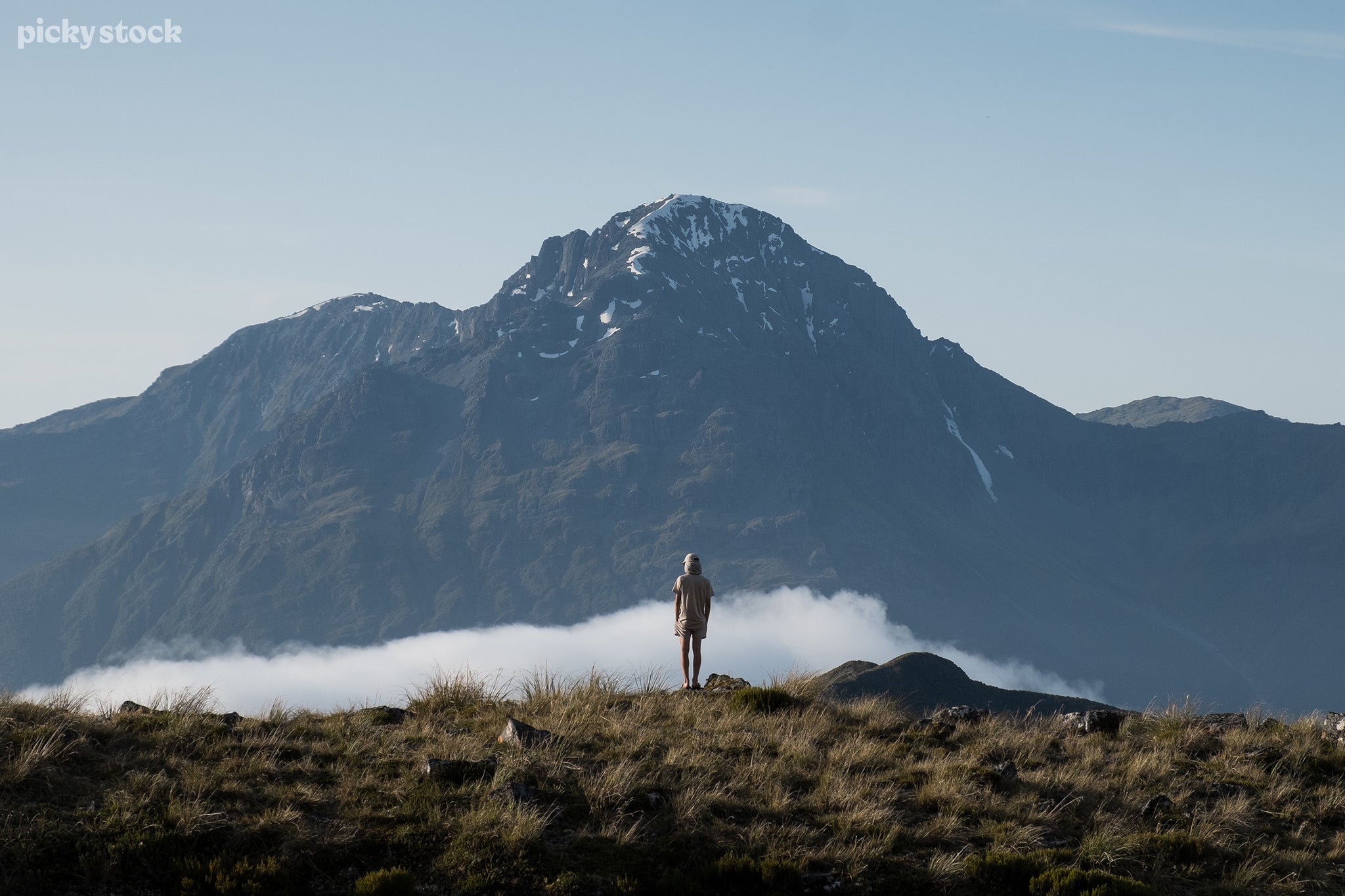 Landscape of a person standing atop a grassy hill overlooking a swell of dense white fog crawling through the valley while a snowy mountain reaches high above overwhelming the ground below in shade.
