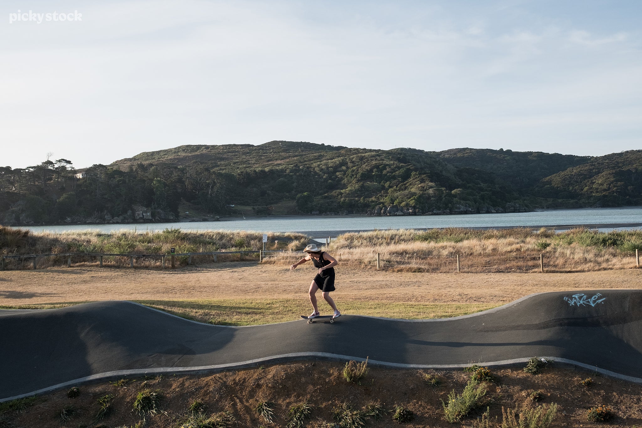 Landscape of a woman in black with pink shoes skateboarding along a skatepark trail surrounded by luscious wild grasses and a wire fence leading towards a body of water.