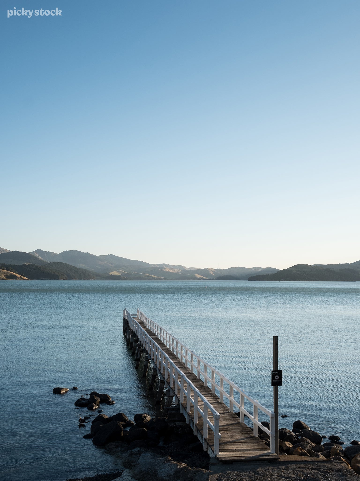 Portrait of a thin  jetty stretching out into the water made of plain wood and white handrails flanked by rocks and a sign saying ‘no animals. Grassy hills can be seen rambling the horizon in the distance.