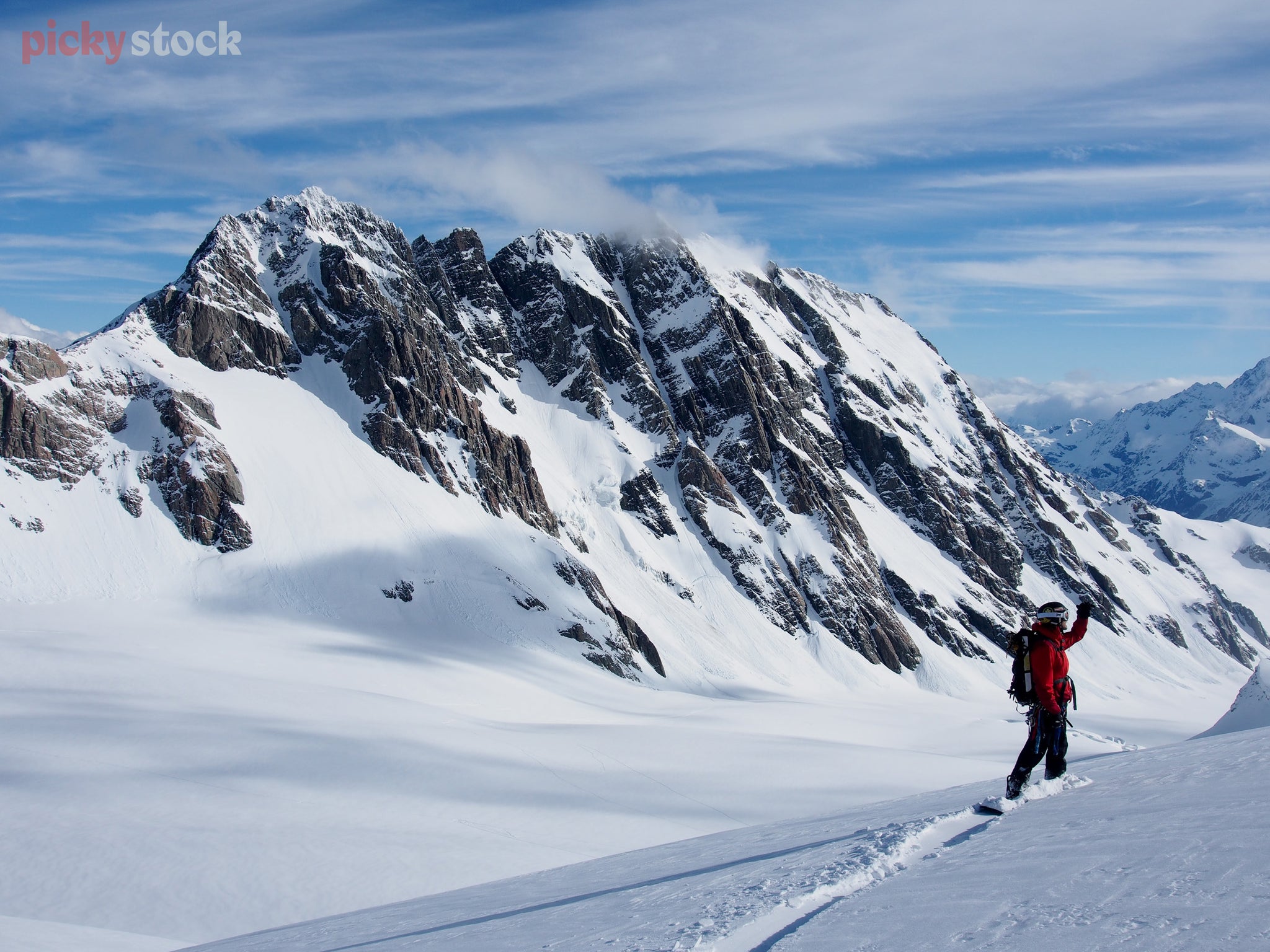 Landscape of a man in a bright red jacket traversing an epic mountain range on a snowboard entrenching neat grooves into the snow as they overlook the slopes.