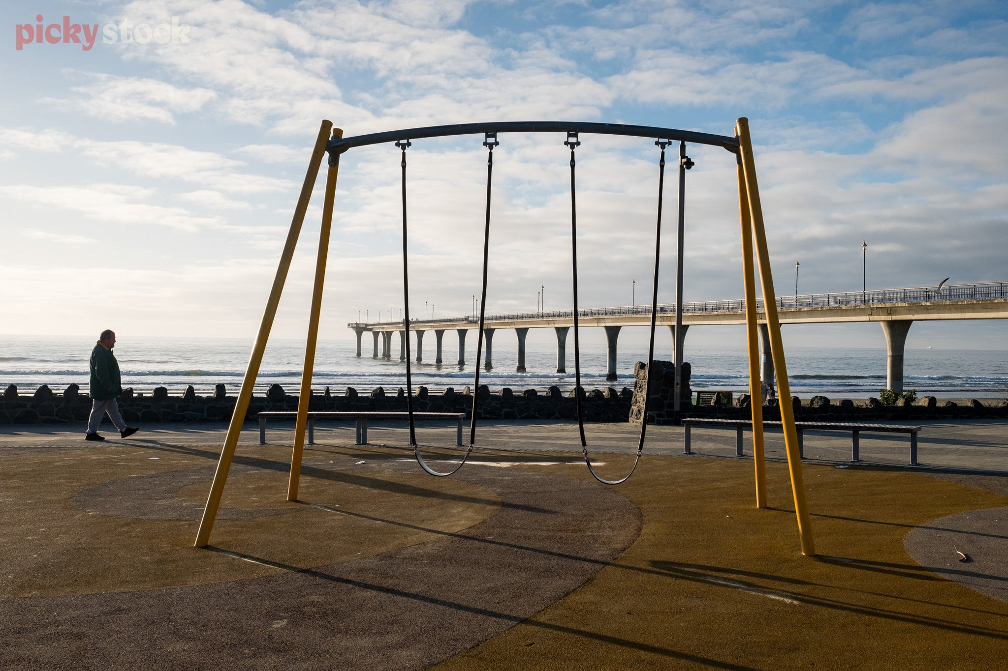 Old yellow double swingset sits empty on the beach front, as an older, unrecognisable gentleman walks past in the background. 