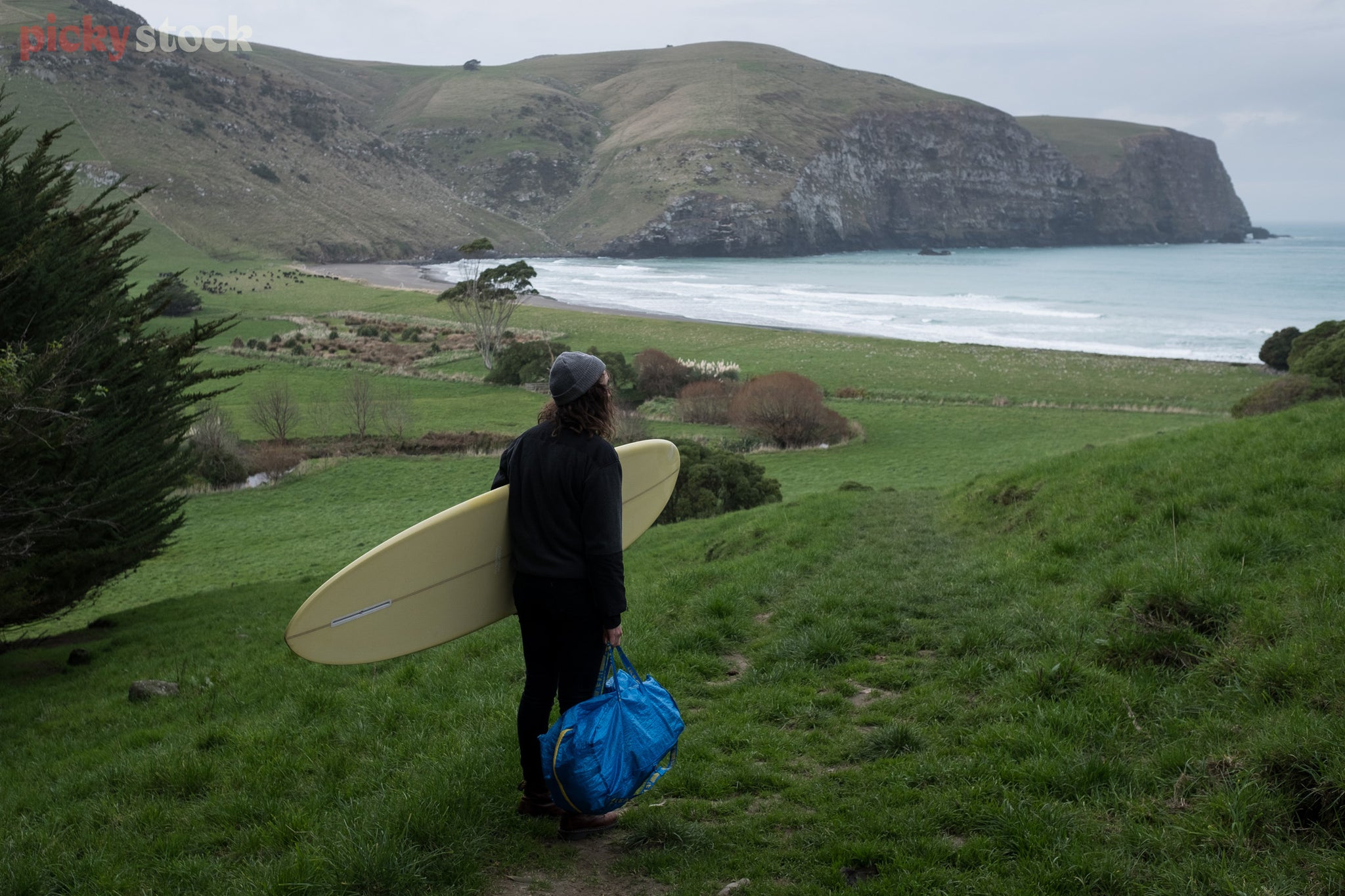 Surfer with beanie, longboard and bag of gear checks out the surf conditions from a hilly track. It's a grey chilly day and he wears long sleeve black top and long dark jeans. 