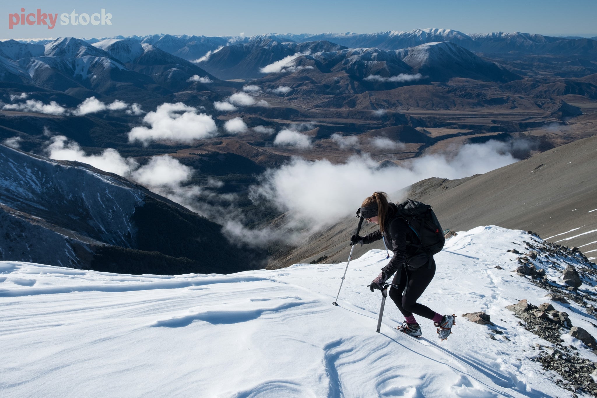 Female tramper uses poles to climb through the snowy-capped mountain ridge, a mountain range can be seen in the background behind. 