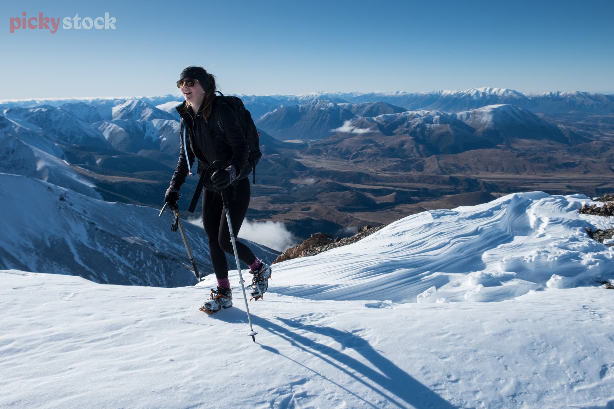 Female tramper uses poles to climb through the snowy-capped mountain ridge, a mountain range can be seen in the background behind. She smiles as she gets to the top. 